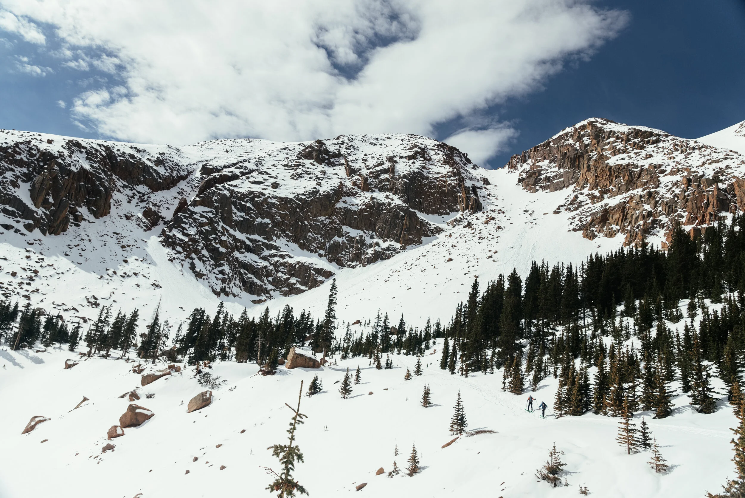 Snow-covered mountain landscape with tall pine trees and two people hiking in the foreground.