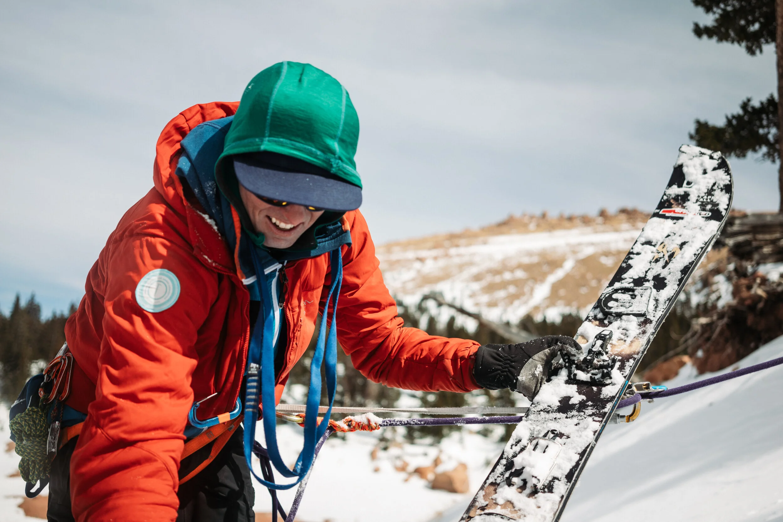 A person dressed in winter gear, including an orange jacket, green hood, and gloves, is adjusting a snowboard on a snowy slope with trees and mountains in the background.