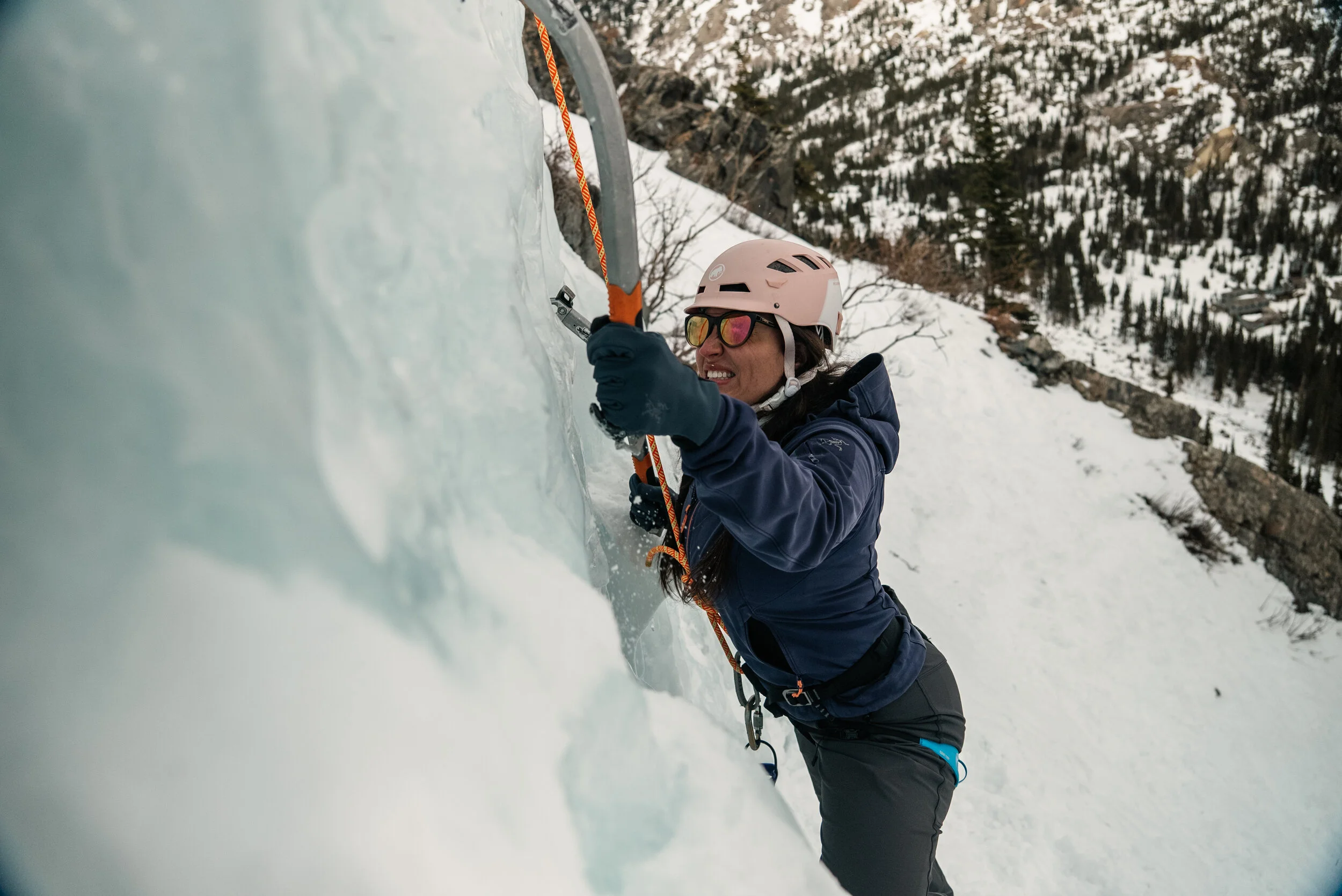 A woman ice climbing on a frozen ice wall outdoors during winter, wearing a pink helmet, sunglasses, a navy jacket, and gloves, using an ice axe to ascend the ice.