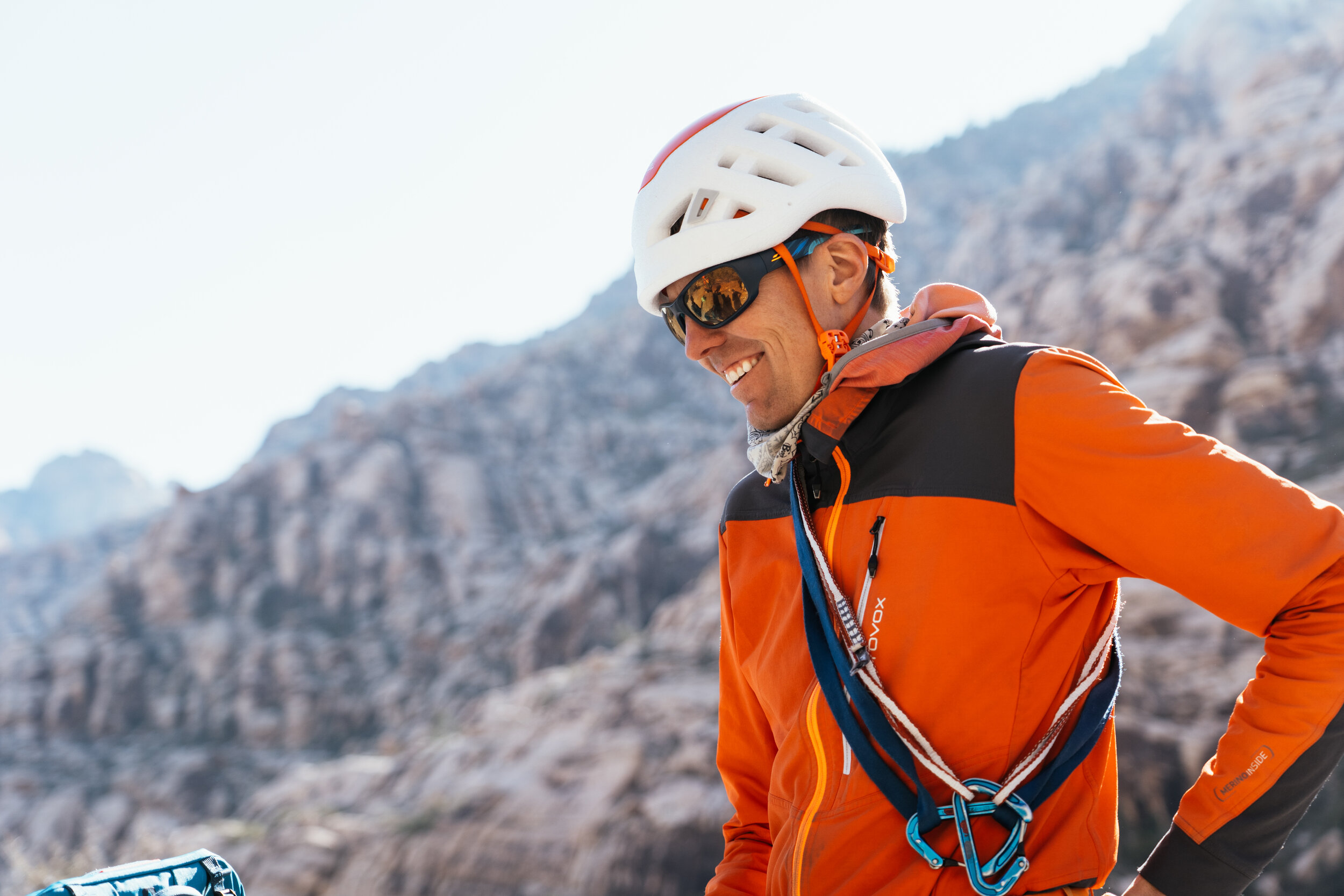 A smiling man wearing a white helmet, sunglasses, an orange and black jacket, and climbing gear stands outdoors with rocky mountains in the background.