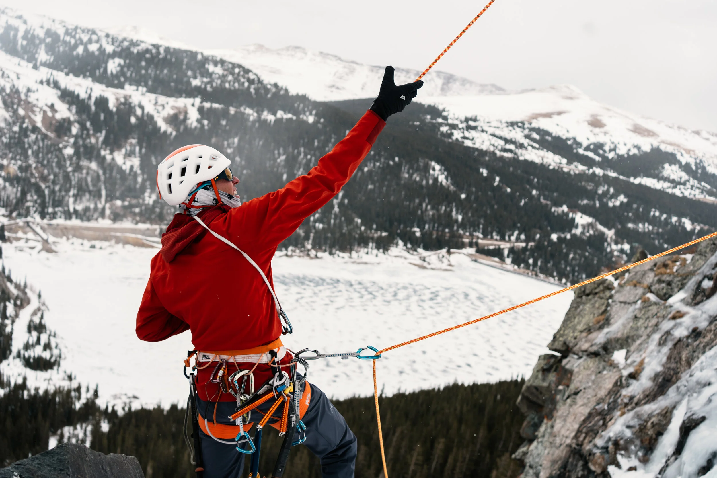 A mountain climber wearing a helmet and a red jacket tied to a safety rope, and reaching out with an ice tool in a snowy mountain landscape.