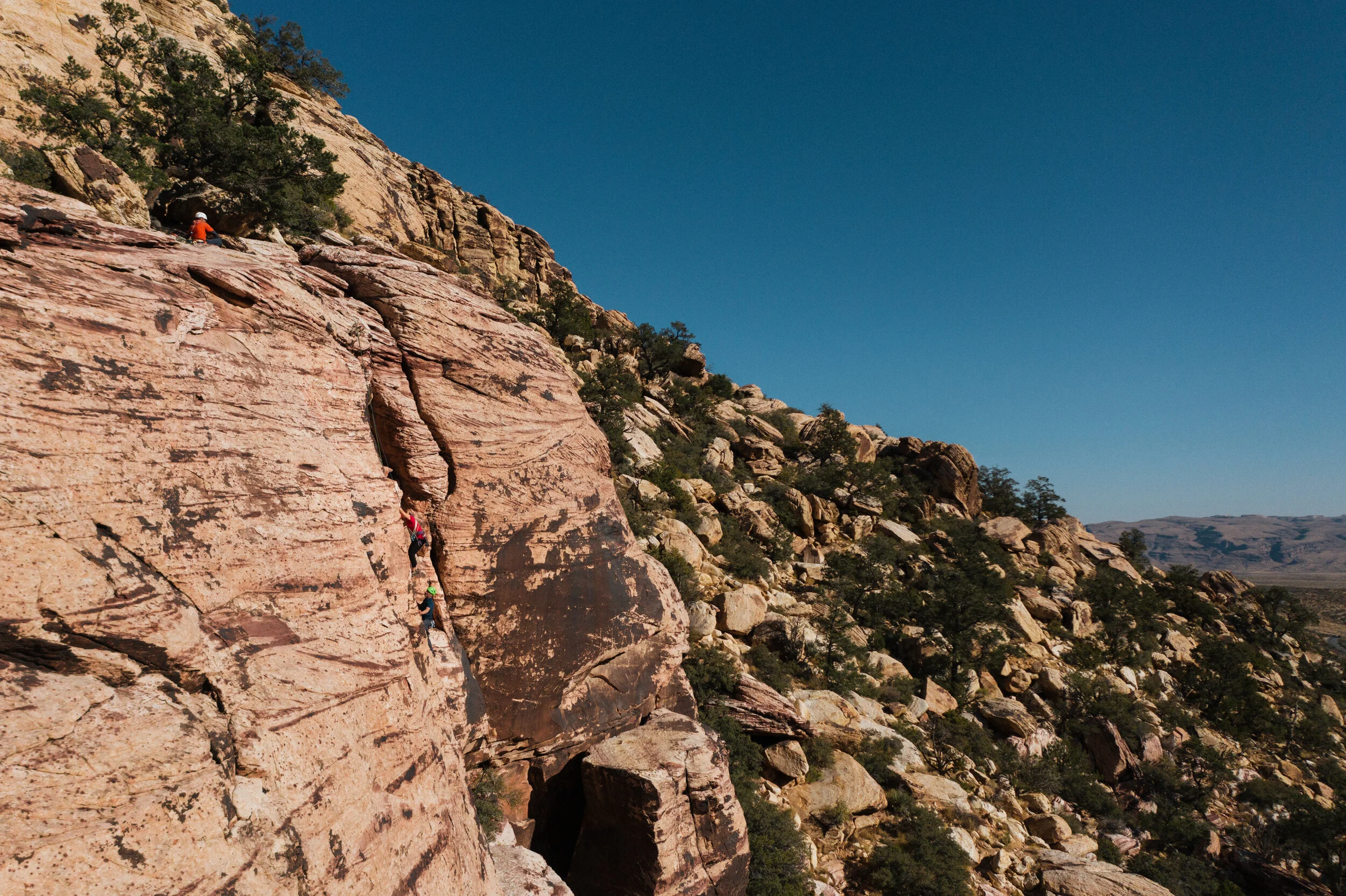 People rock climbing on a large, reddish-brown rock formation with a desert landscape in the background under a clear blue sky.