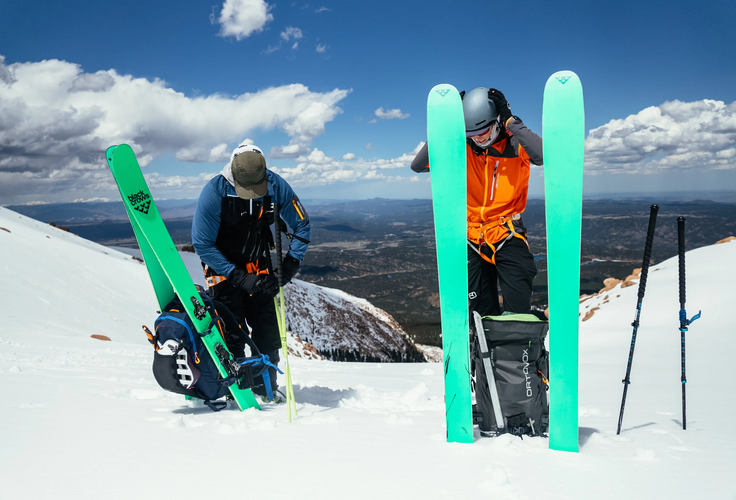 Two people preparing for skiing on a snowy mountain with a view of a valley and mountains in the distance. They have skis, poles, and backpacks.