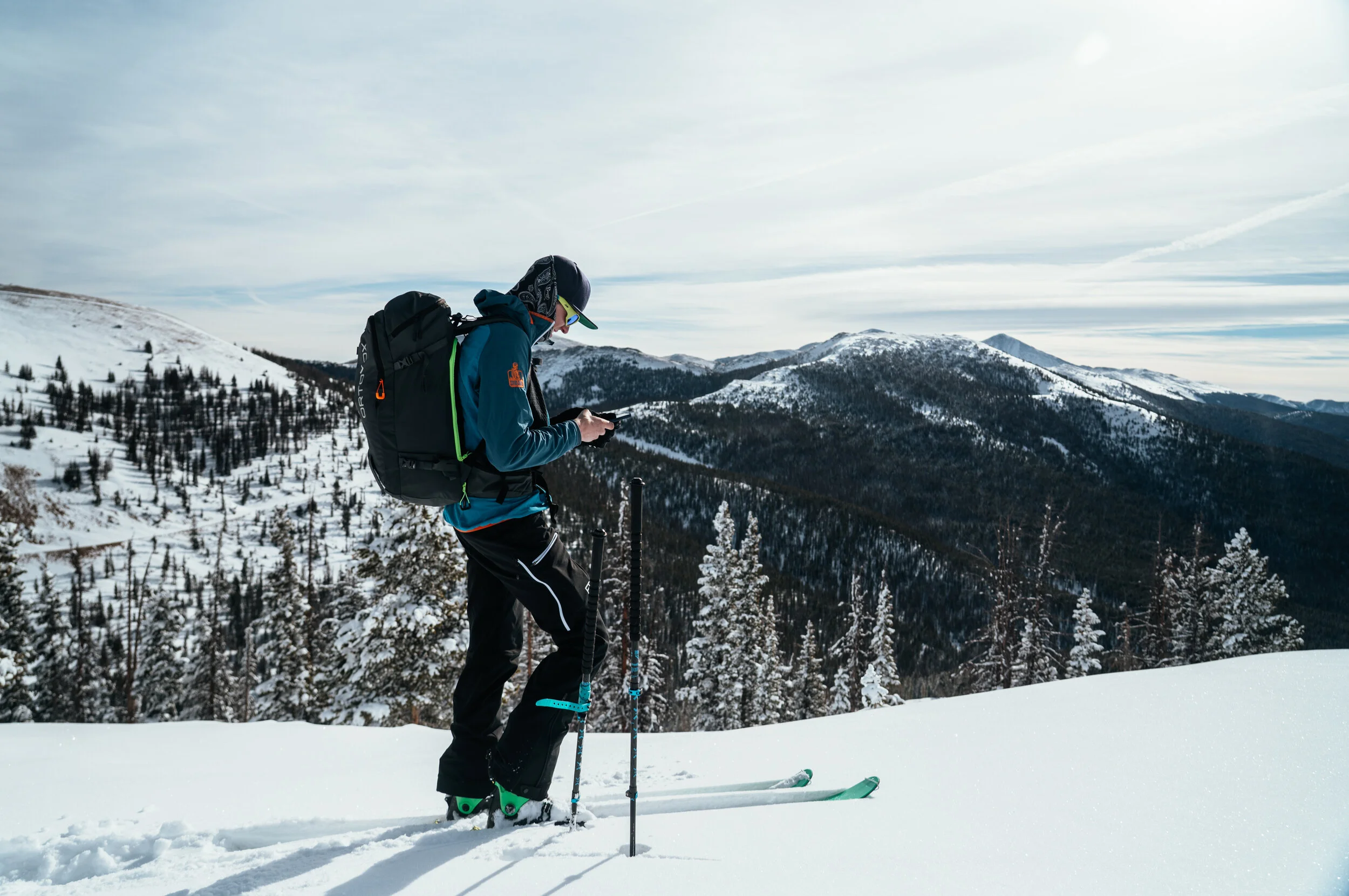 A person in winter gear with skis and poles standing on snow-covered landscape, looking at their phone, with snow-covered mountains and evergreen trees in the background.