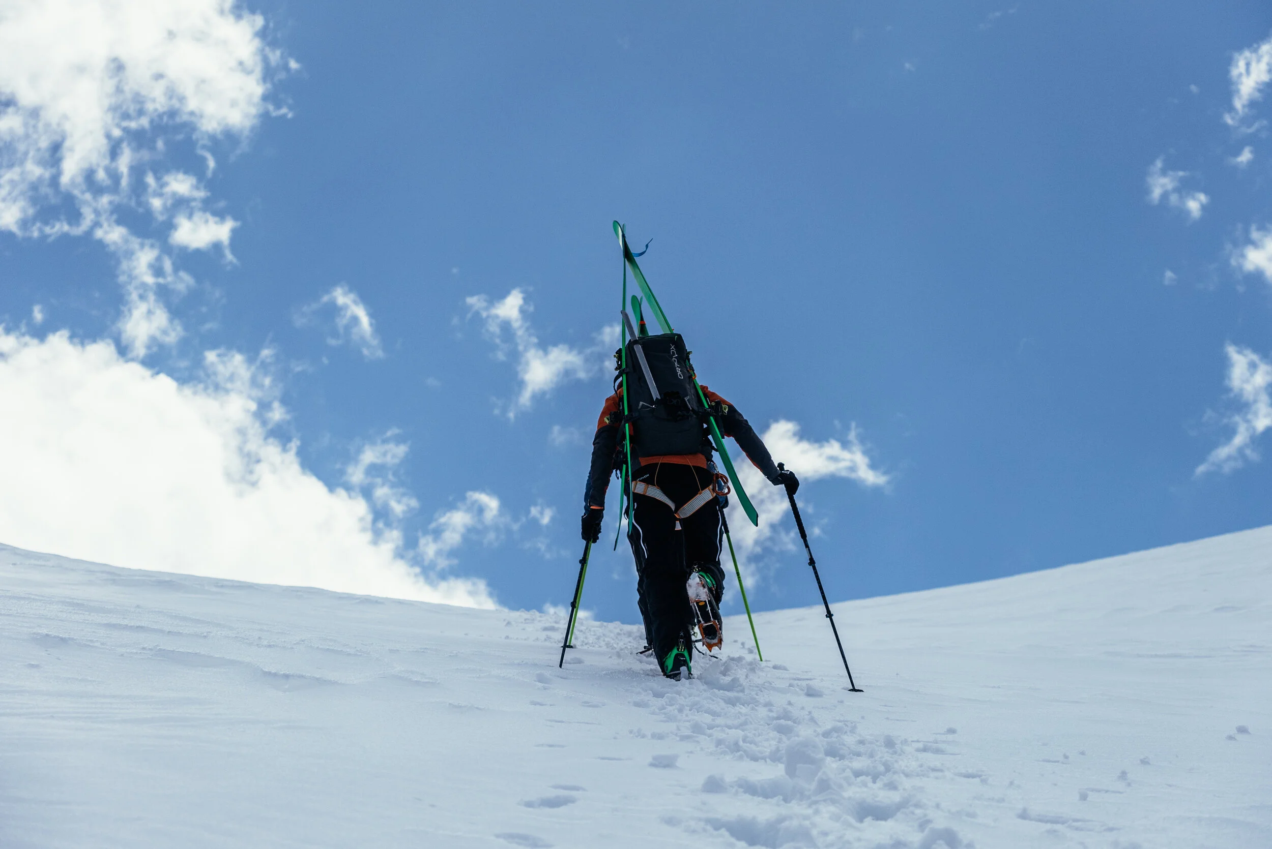 A person dressed in winter gear trekking up a snow-covered mountain slope with ski equipment on their back under a blue sky with clouds.