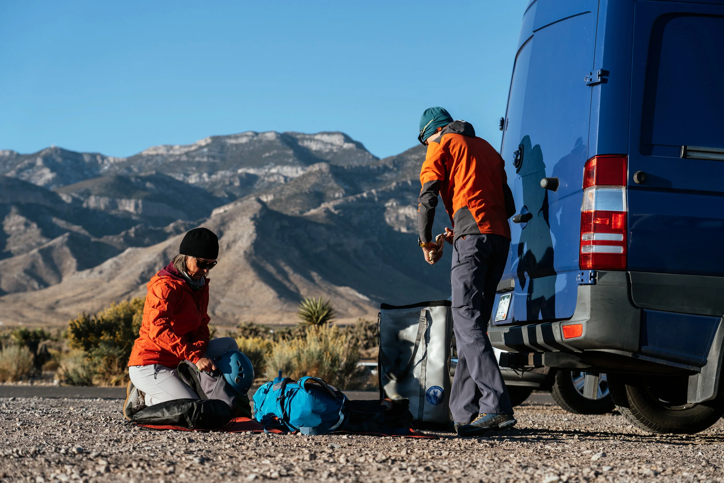 Two people preparing gear beside a blue van on a gravel road with mountains and desert plants in the background.