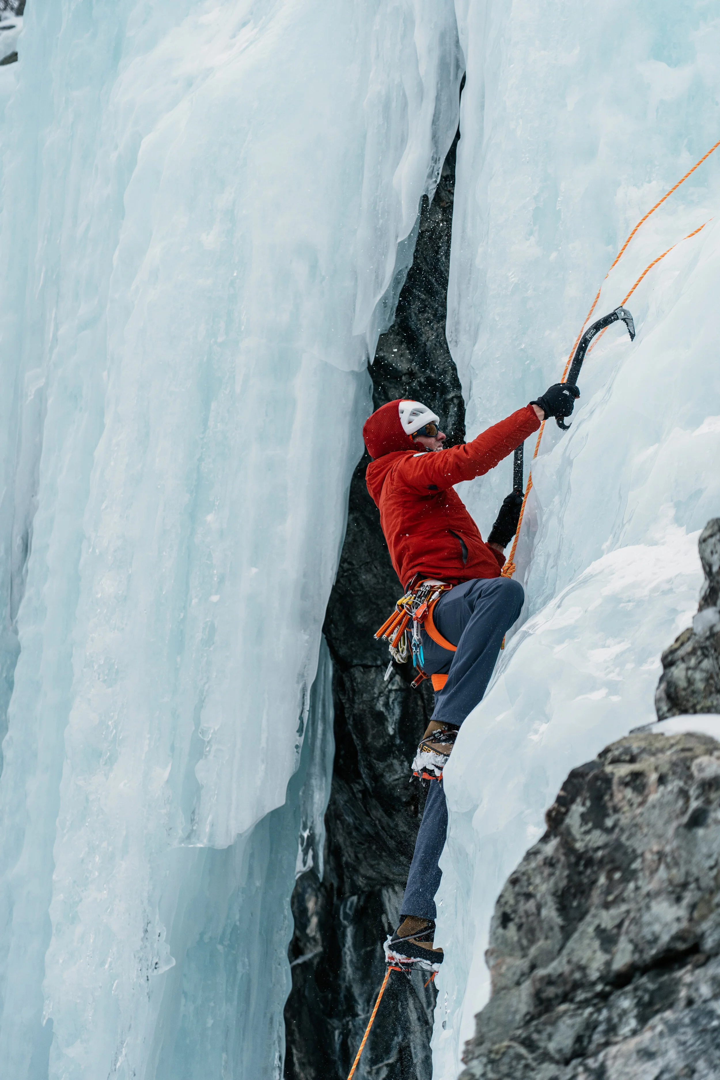 Climber in red jacket and helmet ascending an icy vertical surface with ice axes and ropes.