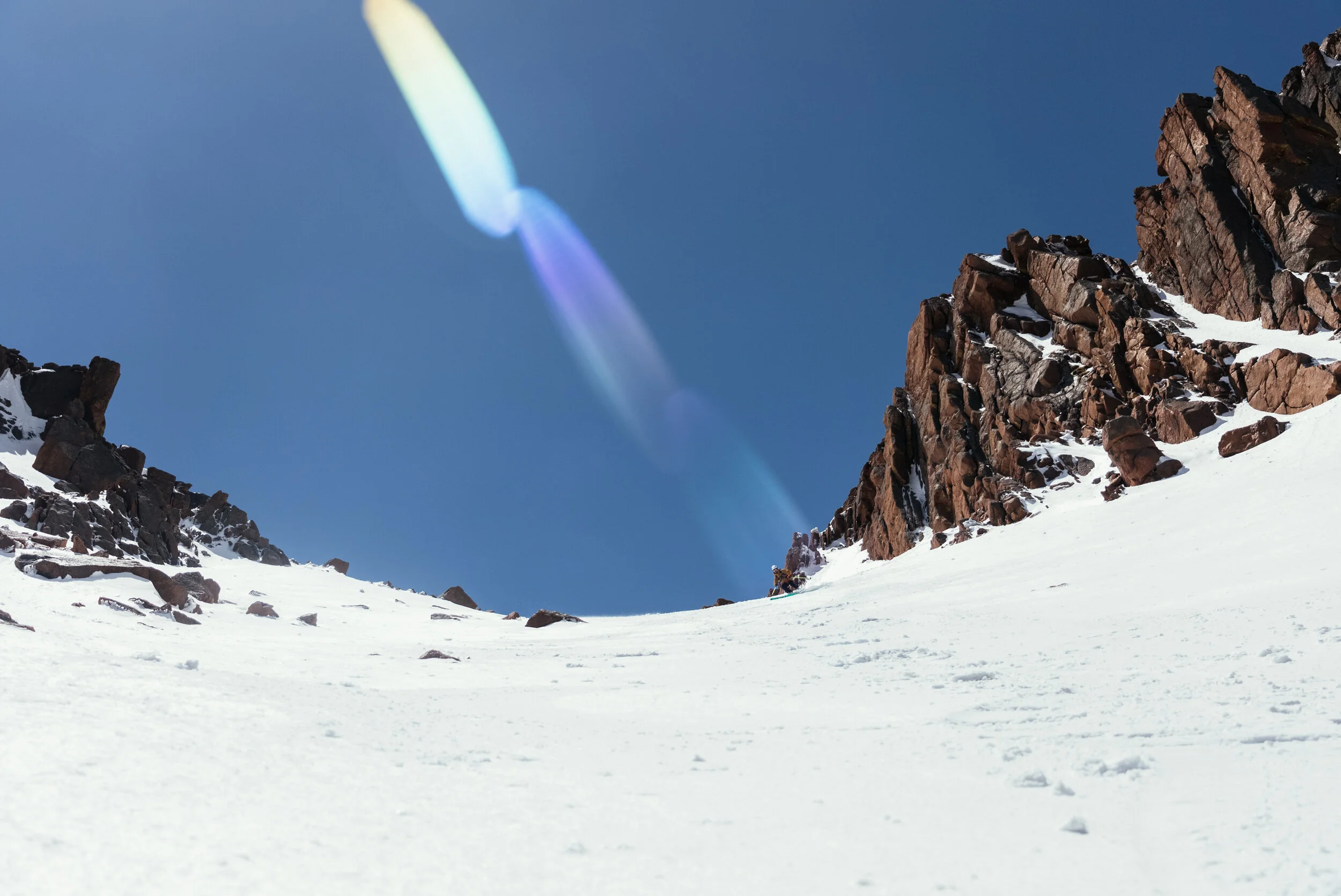 Snow-covered mountain slope with rocky cliffs on the right and a clear blue sky with a rainbow-like lens flare.