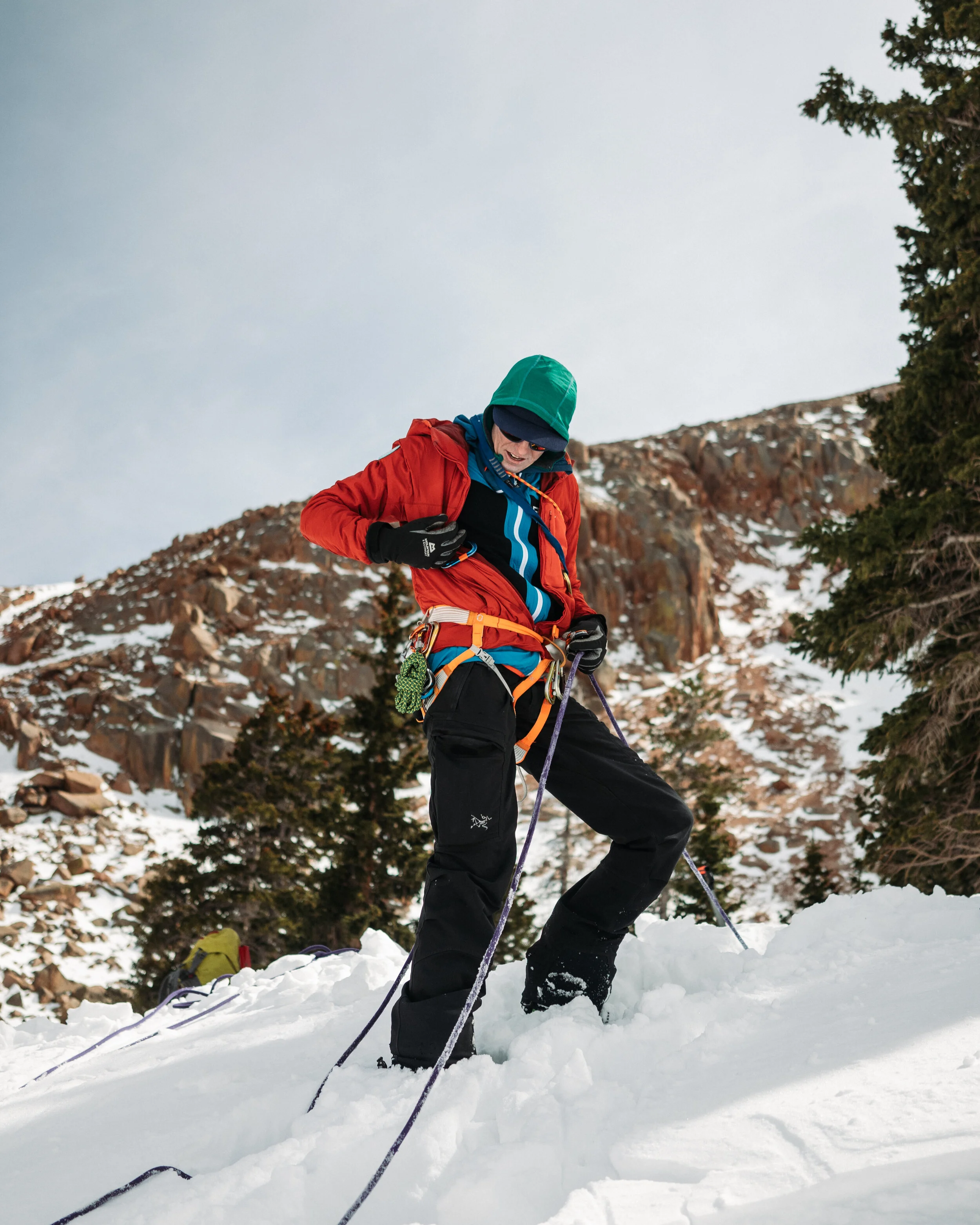 A person dressed in winter gear, including a green hat, red jacket, and black pants, stands in the snow holding ski poles in a snowy mountain landscape with trees and rocky slopes.