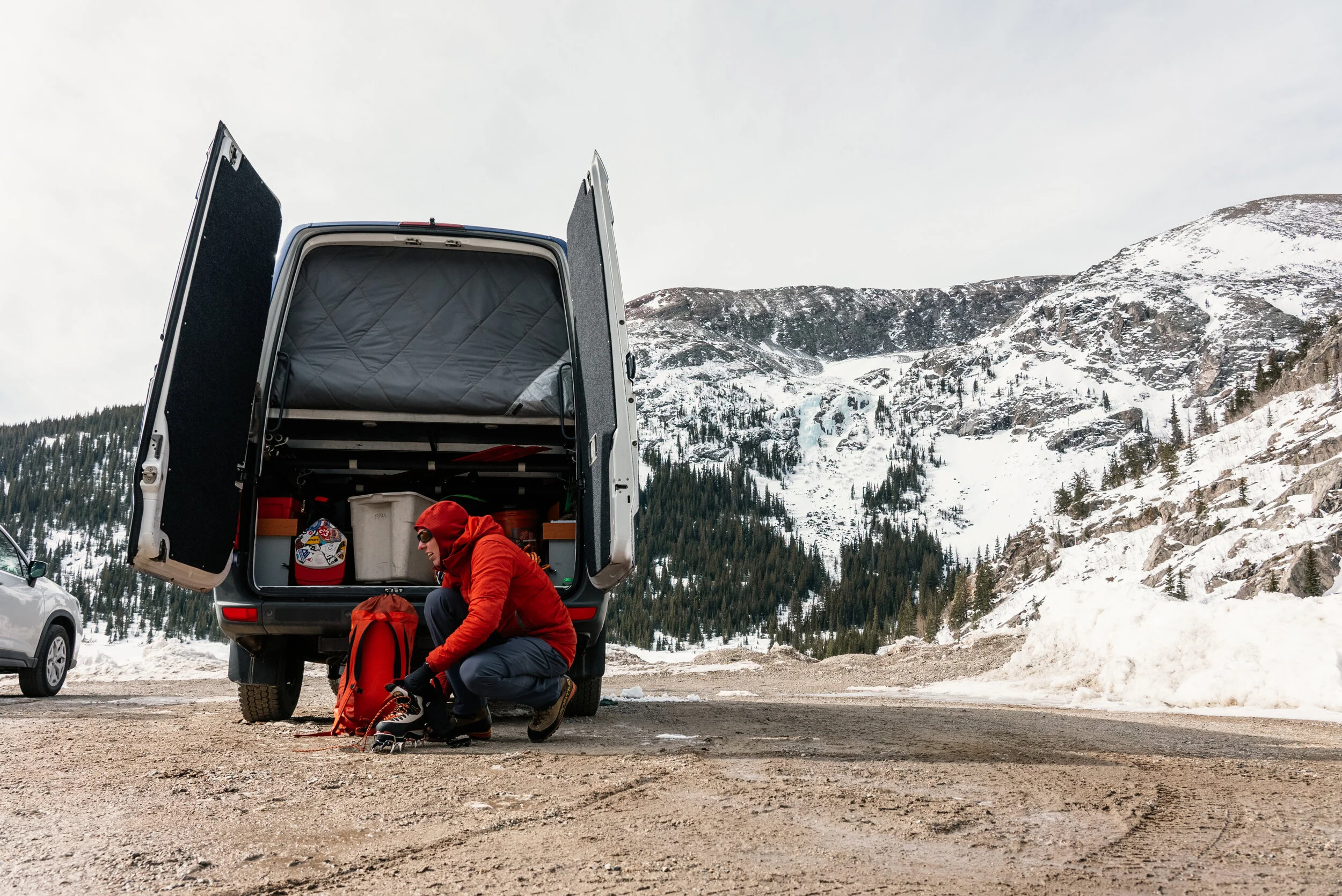 A person in red winter clothing and a red beanie putting on snowshoes beside a van in a snowy mountainous area.