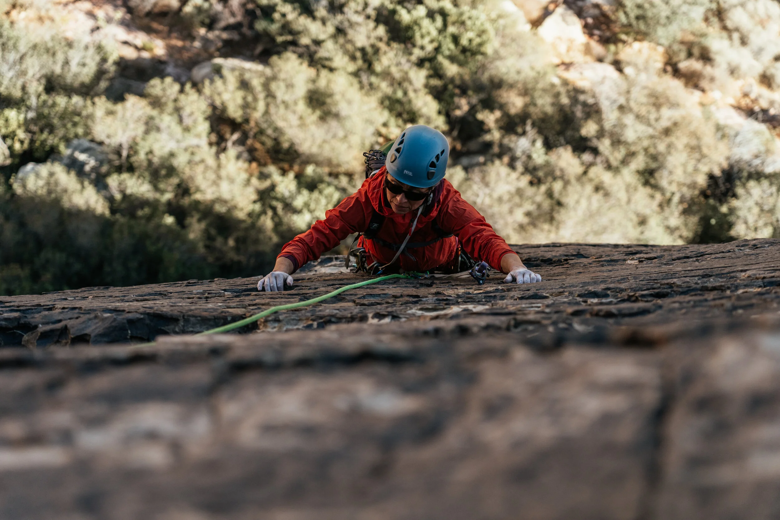 Climber wearing a blue helmet and red jacket, ascending a steep rock face, secured with a climbing rope, against a backdrop of trees and rocks.