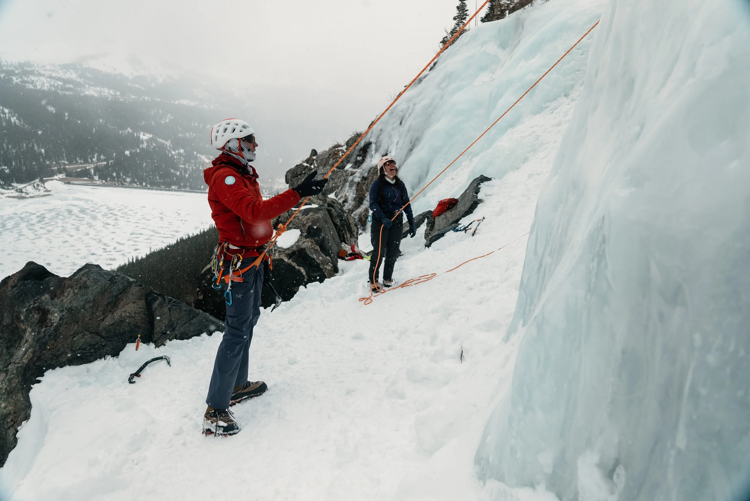 Two climbers in winter gear on a snowy mountain slope, one using an ice axe and the other with climbing ropes, near a large ice formation.