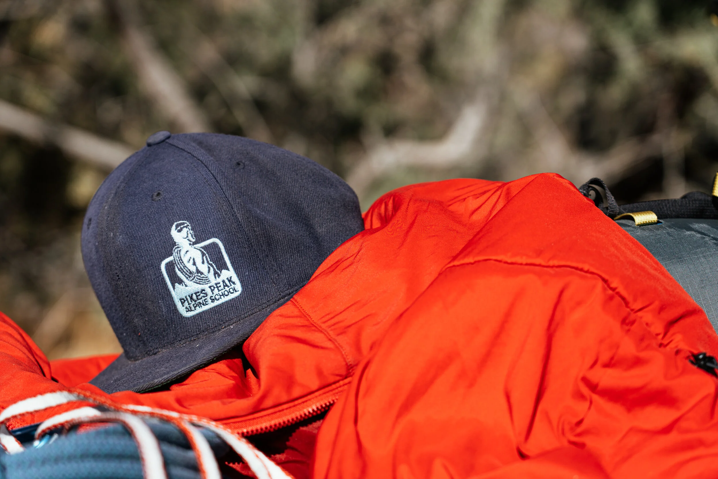 A navy blue baseball cap with a logo of a mountain climber and text 'Pikes Peak Alpine School' on it, placed on top of a red outdoor jacket, with a blurred background of trees.