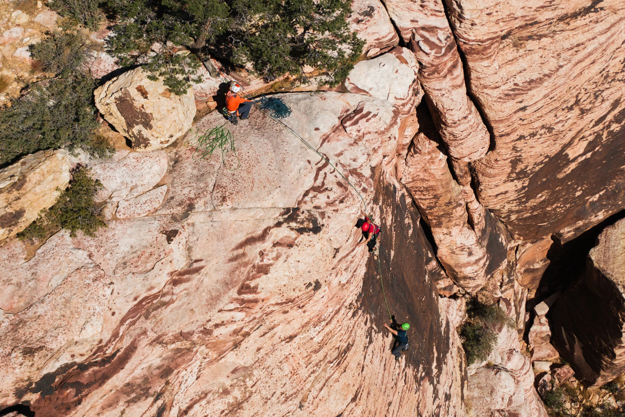 Three rock climbers on a steep, orange and brown cliff face, with one climber securing the others, while a person at the top manages the climbing rope.