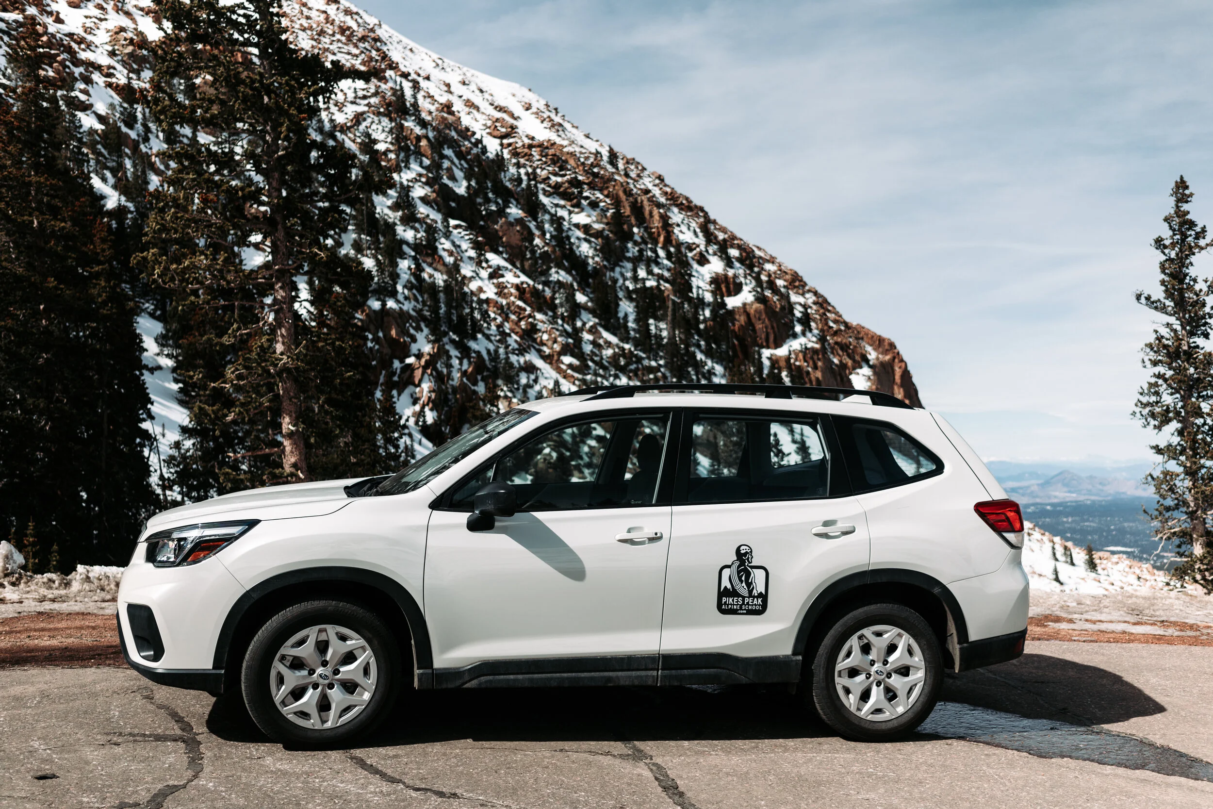 White SUV parked on a mountain road with snow-covered trees and rocky mountains in the background. The vehicle has a logo on the side that reads 'Pikes Peak Alpine School' with a mountain climber icon.