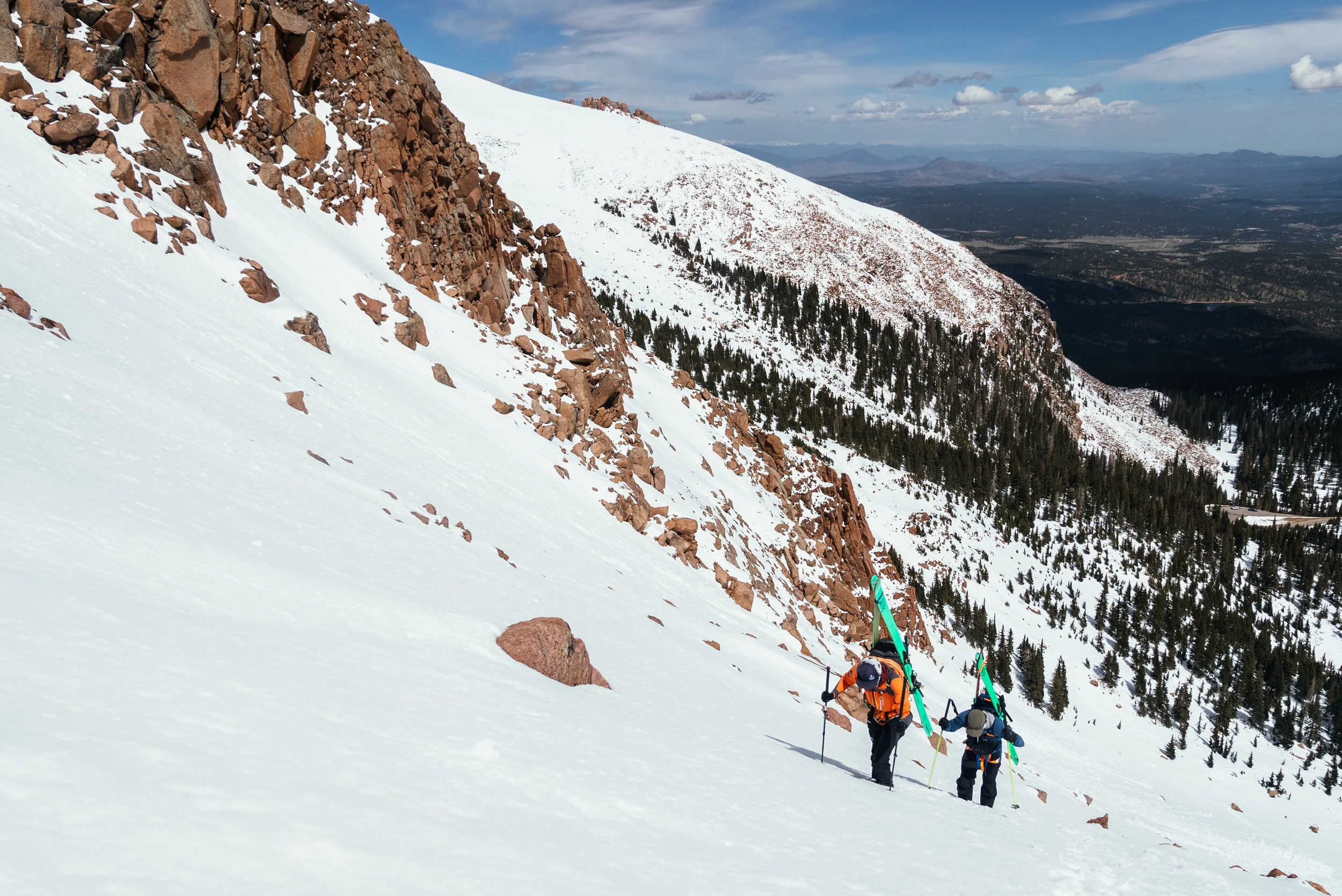 Two hikers in winter gear ascending a snowy mountain slope with skis strapped to their backpacks, surrounded by rocky terrain and distant trees under a partly cloudy sky.