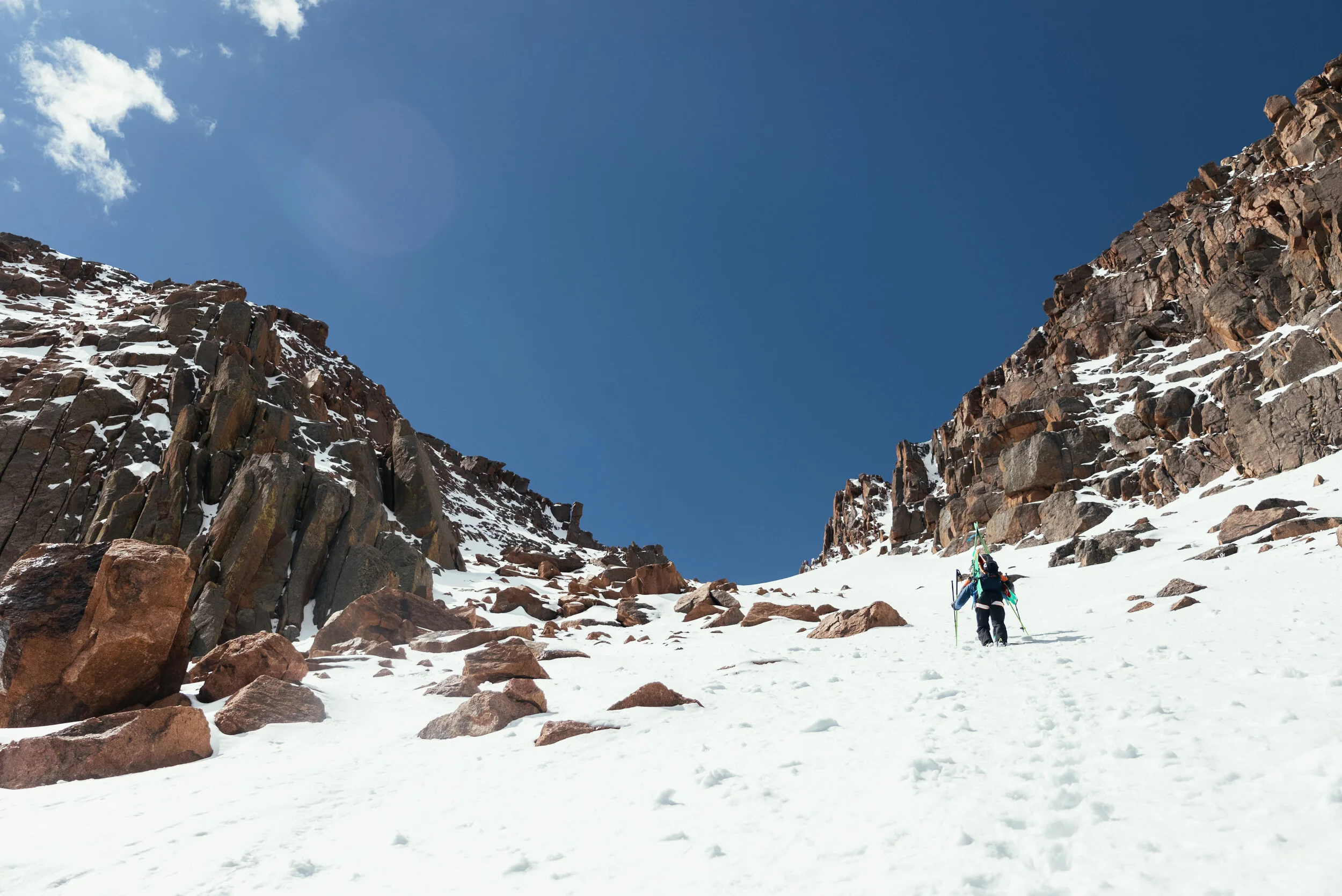 A person hiking up a snowy mountain slope between rocky cliffs under a clear blue sky.
