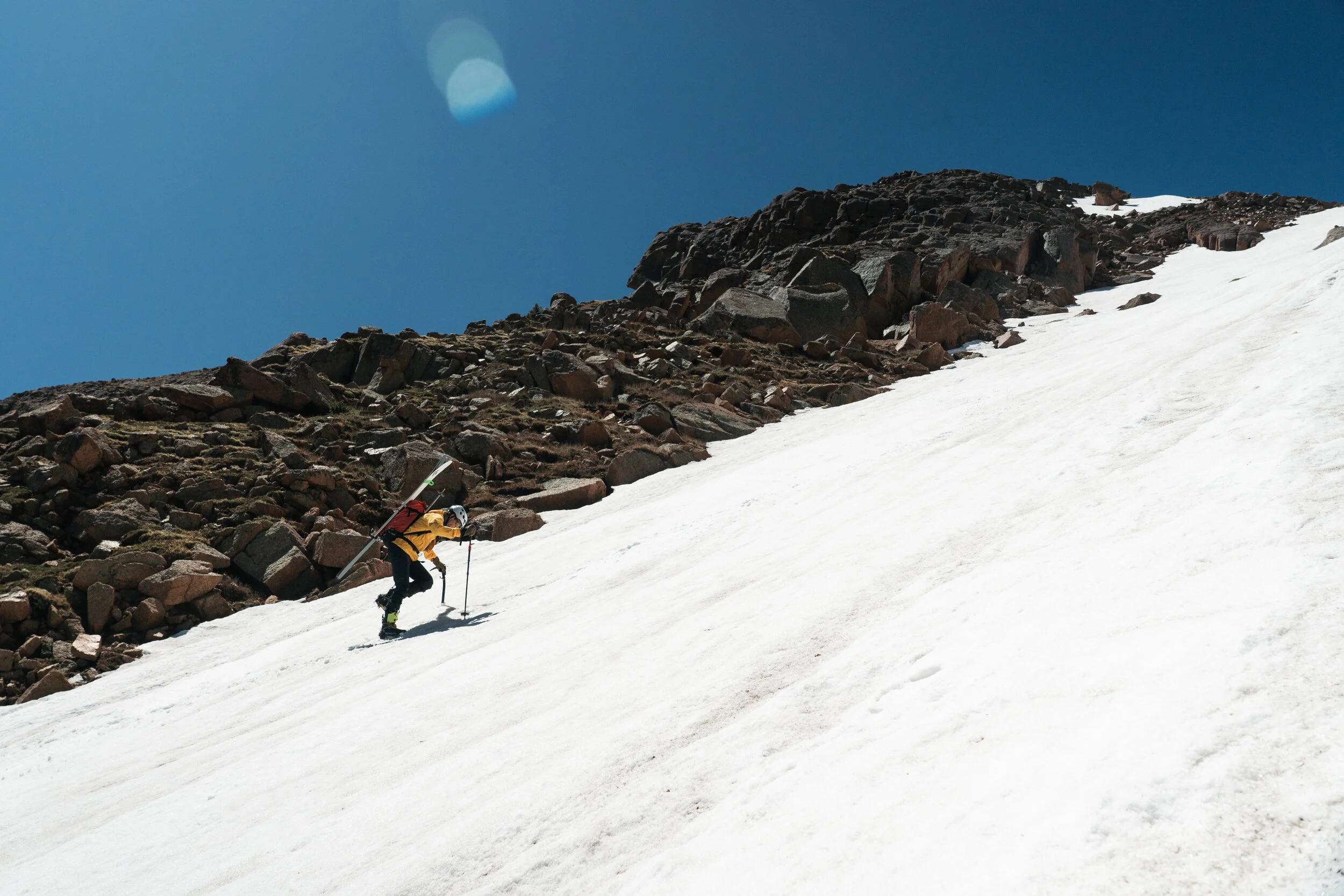 A person in yellow and black winter gear climbing a snowy mountain slope with rocks in the background and a clear blue sky.
