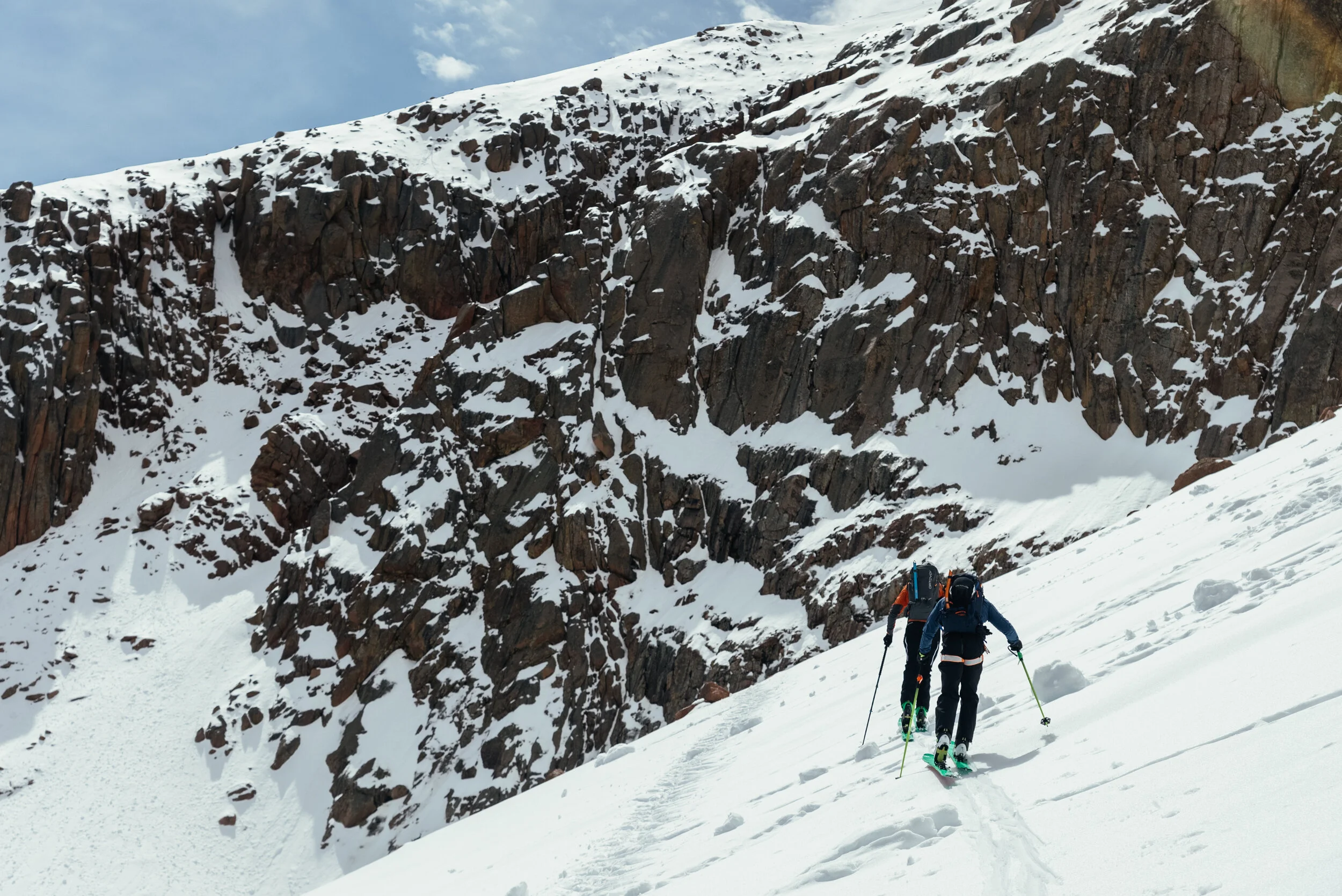 Two hikers wearing backpacks and snow gear ascending a snowy mountain slope with rocky cliffs in the background.