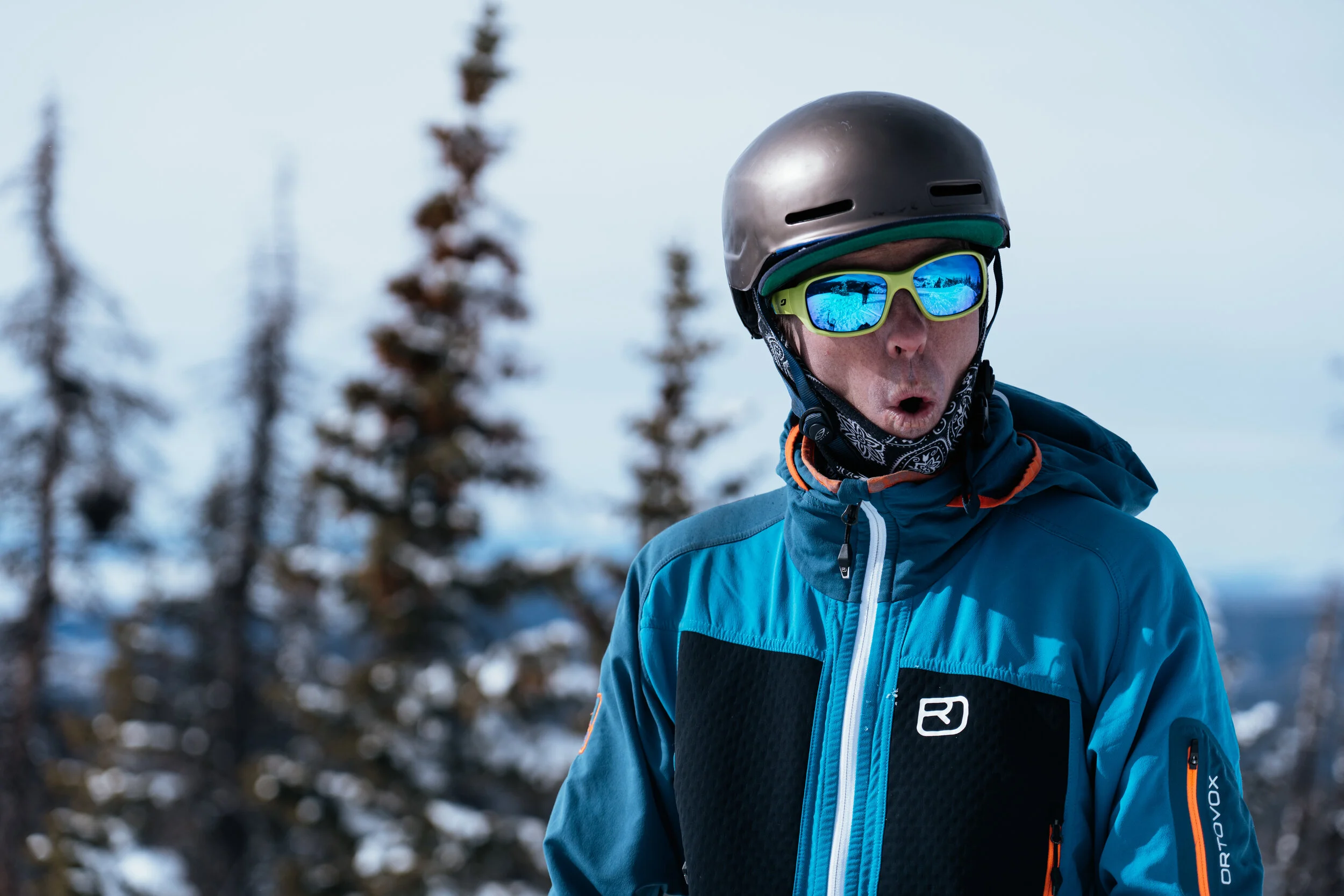A man wearing a helmet, sunglasses, and a blue winter jacket outdoors with snow and trees in the background.