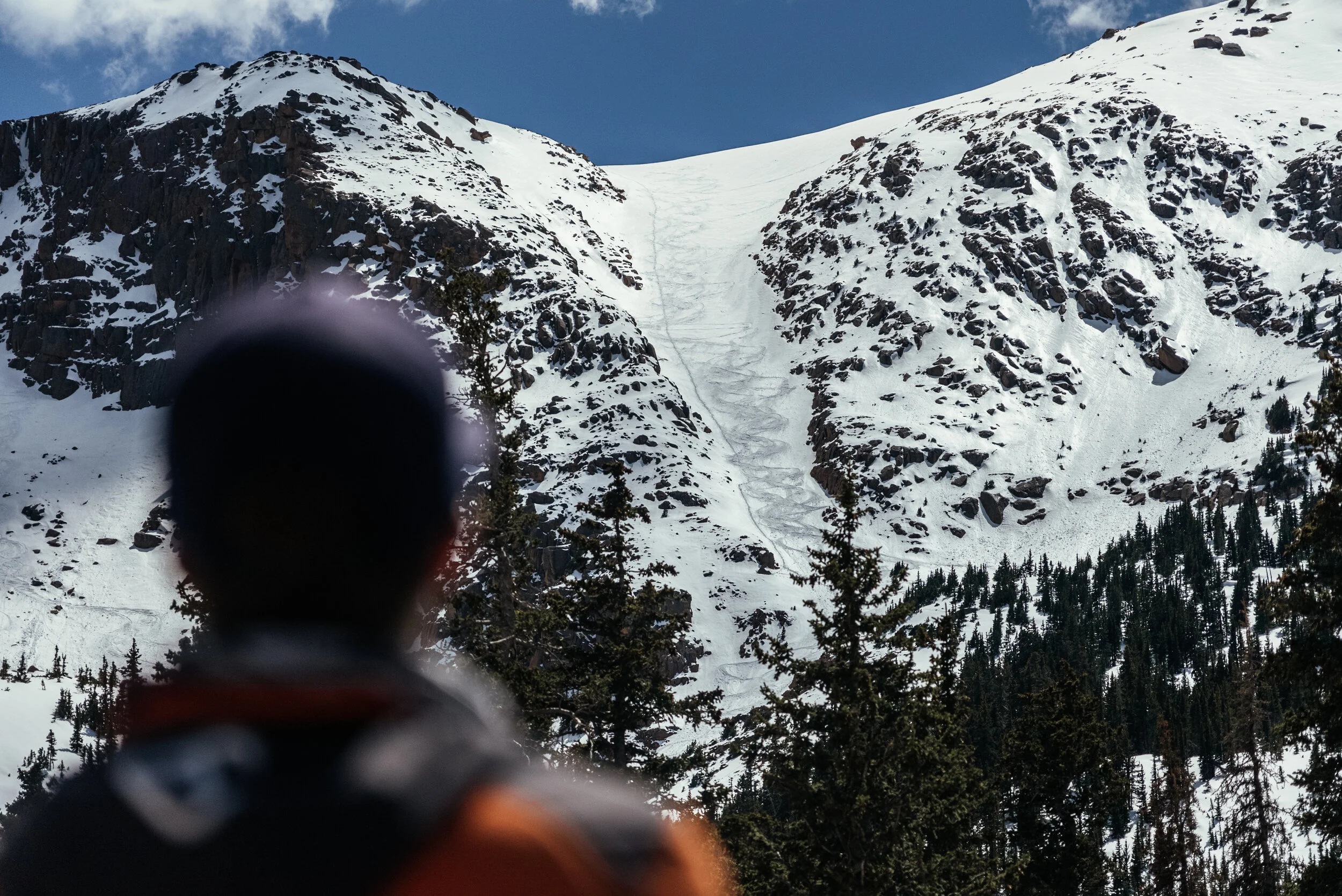 The image shows a person in the foreground with a blurred face, looking towards a snowy mountain with a narrow snow chute or ski run down its face, surrounded by pine trees.