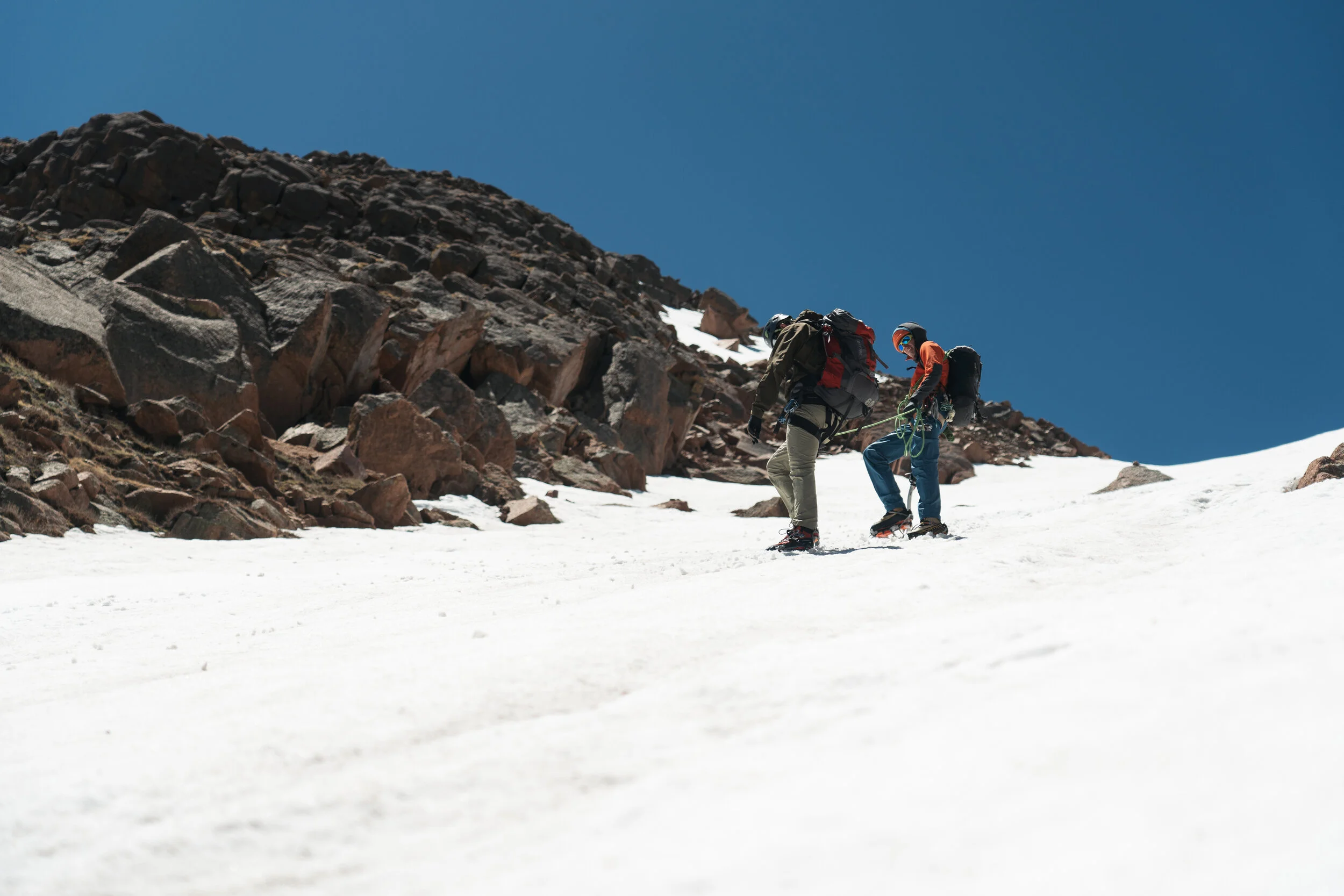 Two climbers in winter gear ascending snow-covered mountainous terrain under a clear blue sky.