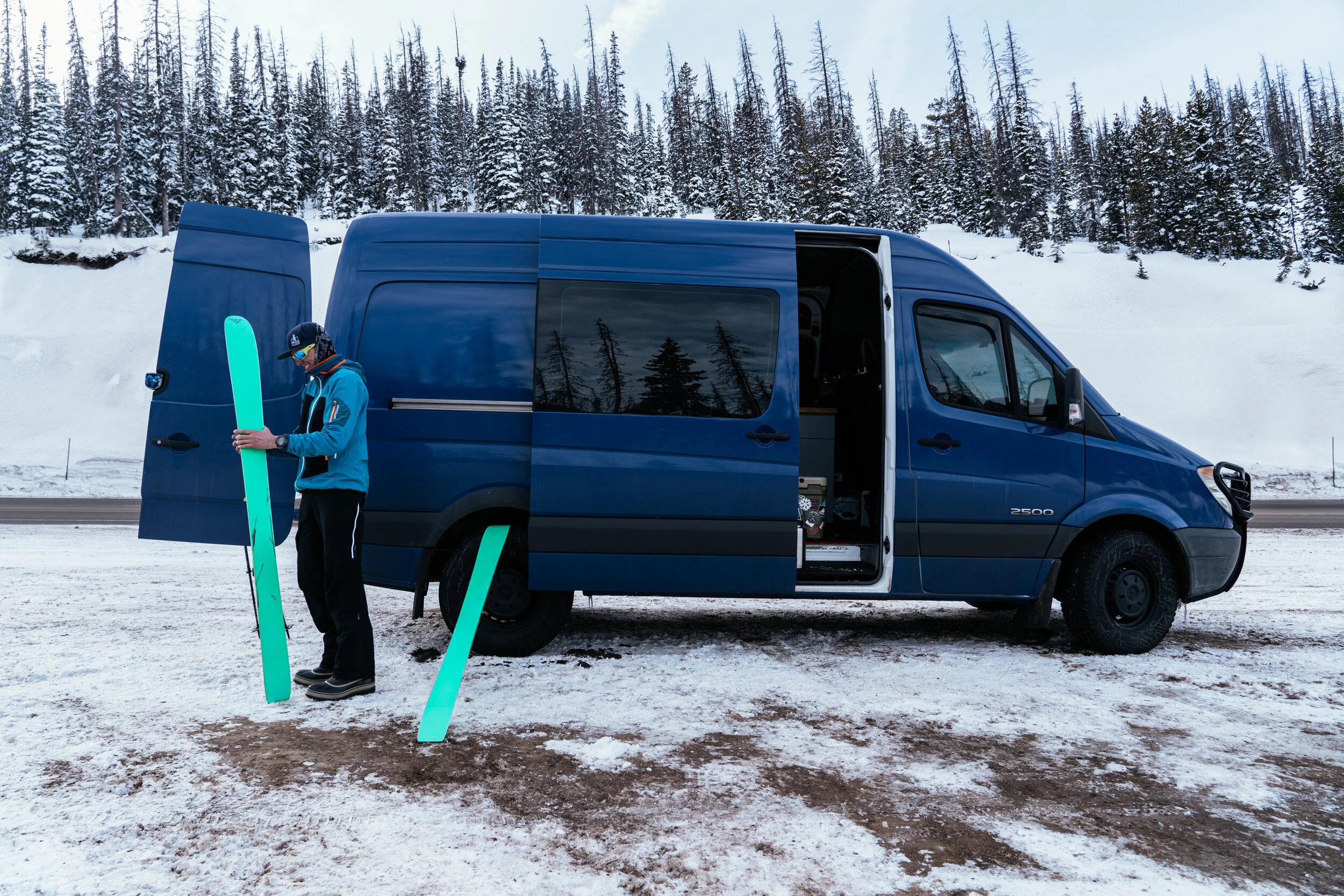 A person dressed in winter sports gear standing next to a blue van on a snowy ground, holding a bright turquoise skis.
