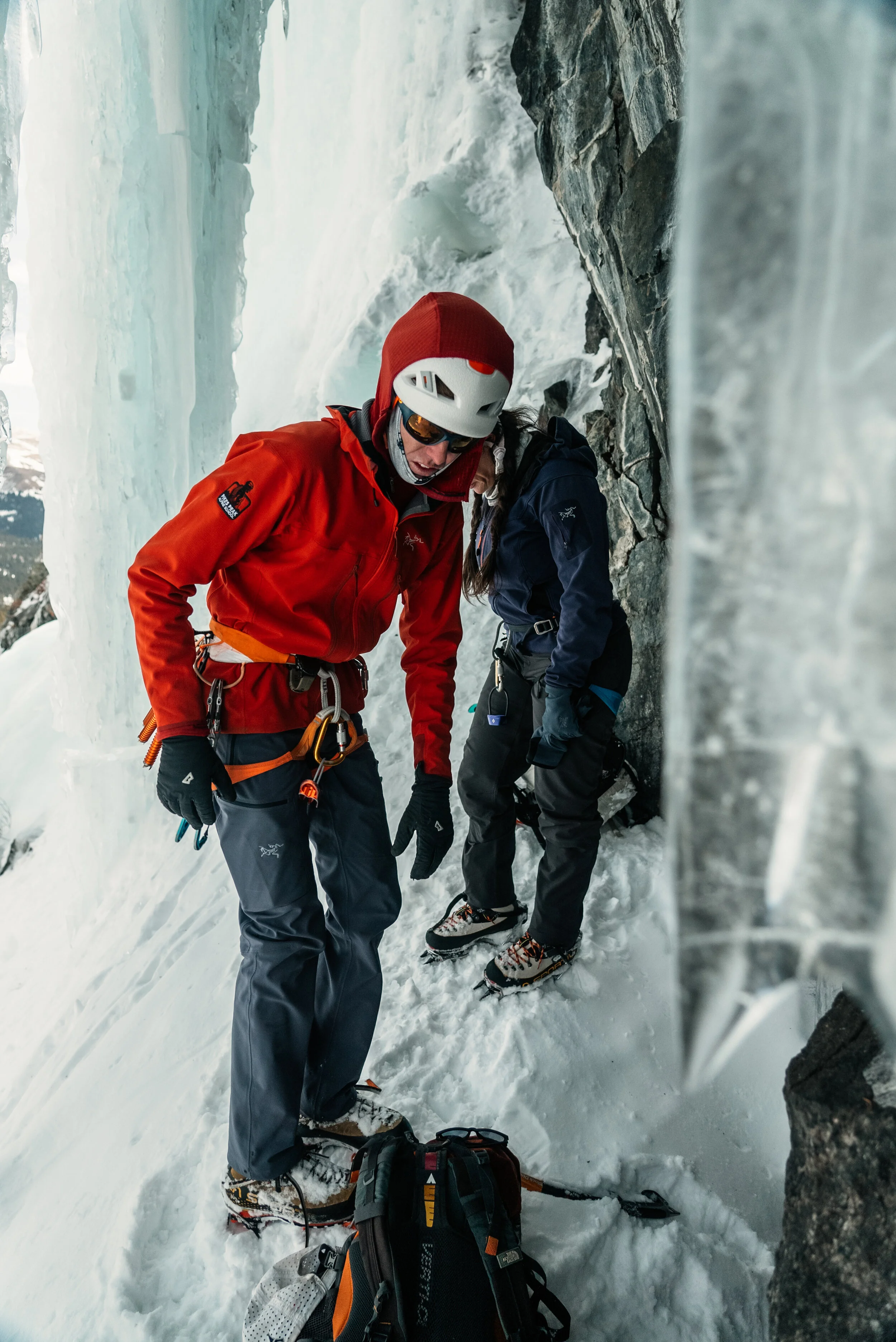 Two climbers in winter gear on an icy mountain slope, with one adjusting equipment near the rock face and the other looking down, surrounded by snow and ice.