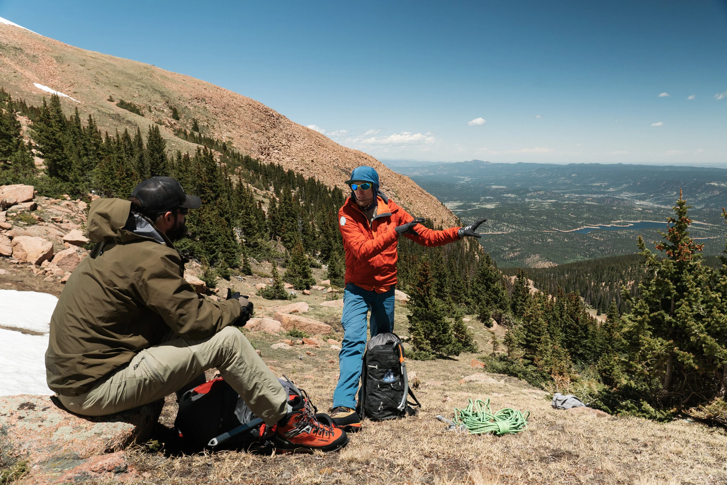Two hikers on a mountain trail, one sitting and the other standing, with a scenic view of a forested valley and mountains in the background.