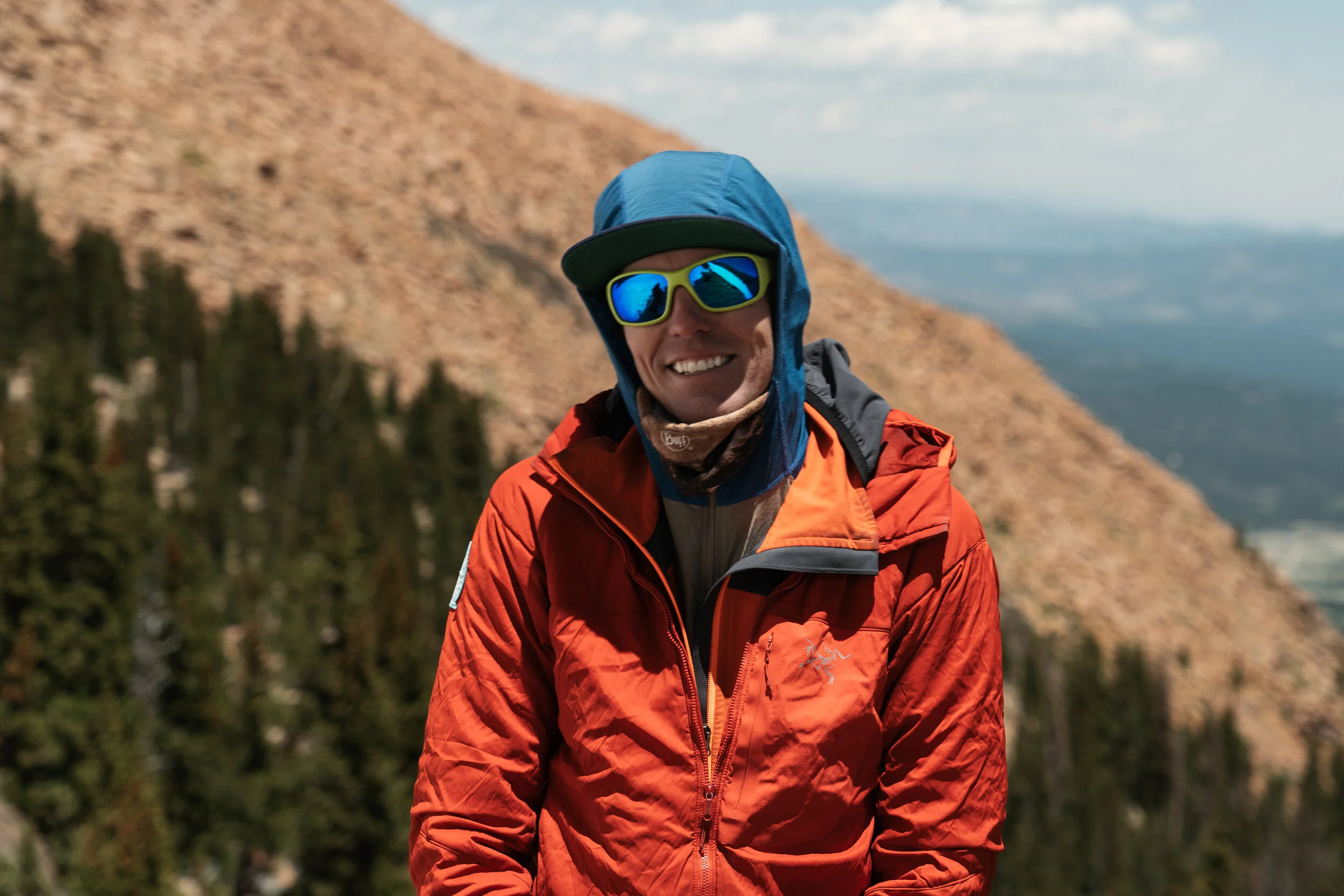 A man wearing a blue hooded jacket, sports sunglasses, and an orange jacket, smiling outdoors with mountains and trees in the background.