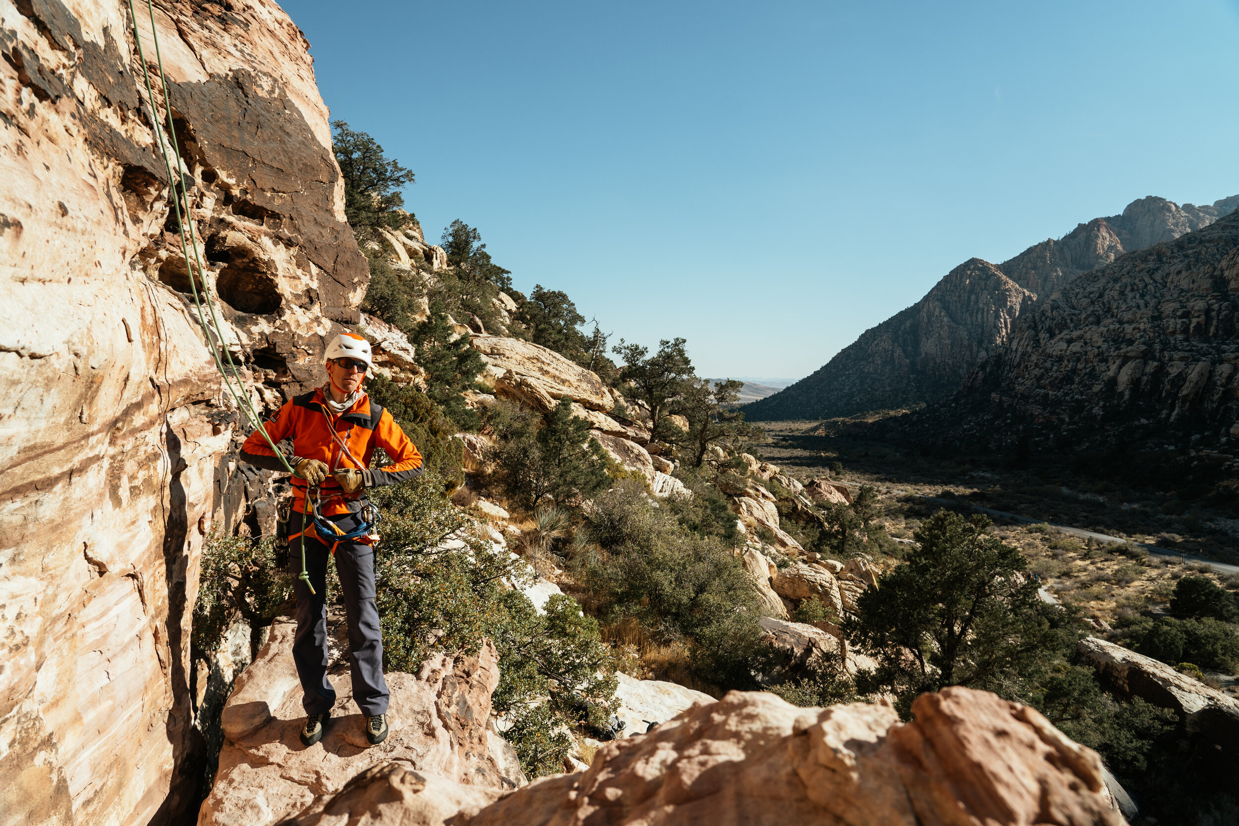 A person in an orange jacket, climbing harness, helmet, and sunglasses, setting up or adjusting rock climbing gear on a rocky hillside with mountainous terrain and sparse vegetation under a clear sky.