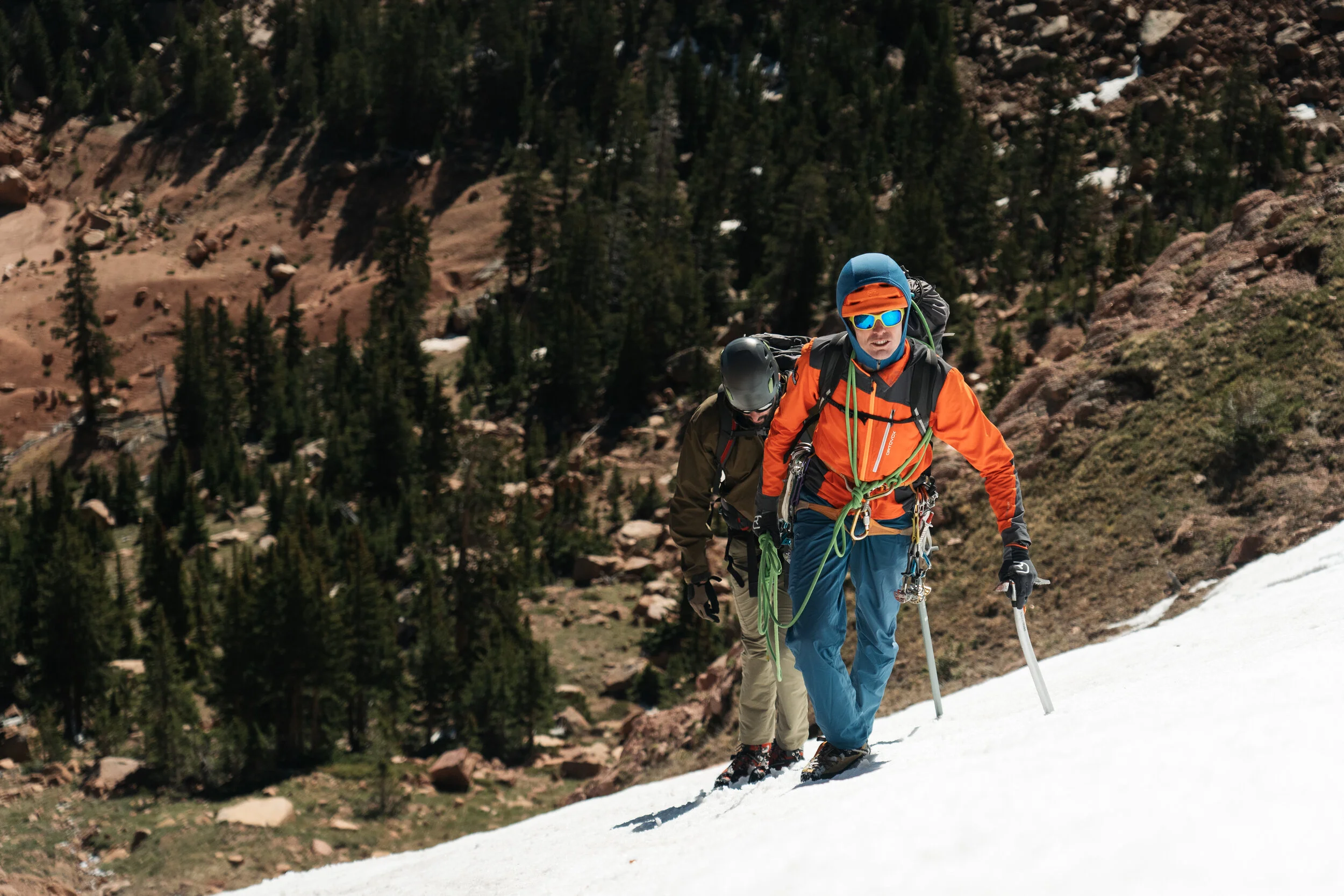 Two climbers ascending a snow-covered mountain slope with a forested mountain landscape in the background.
