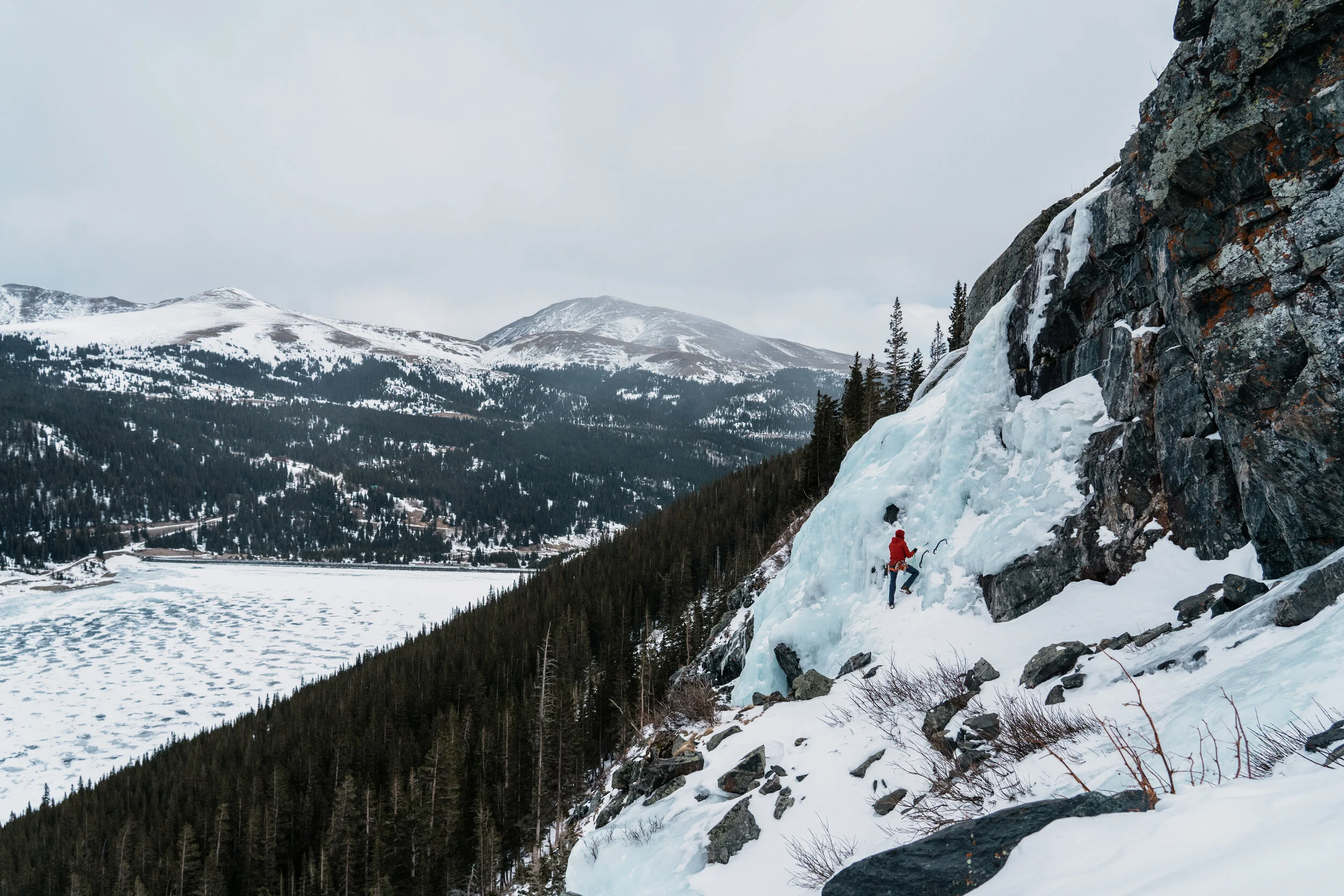 A person in red winter gear climbing an icy waterfall on a mountain slope, with snow-covered mountains and a partially frozen lake in the background.