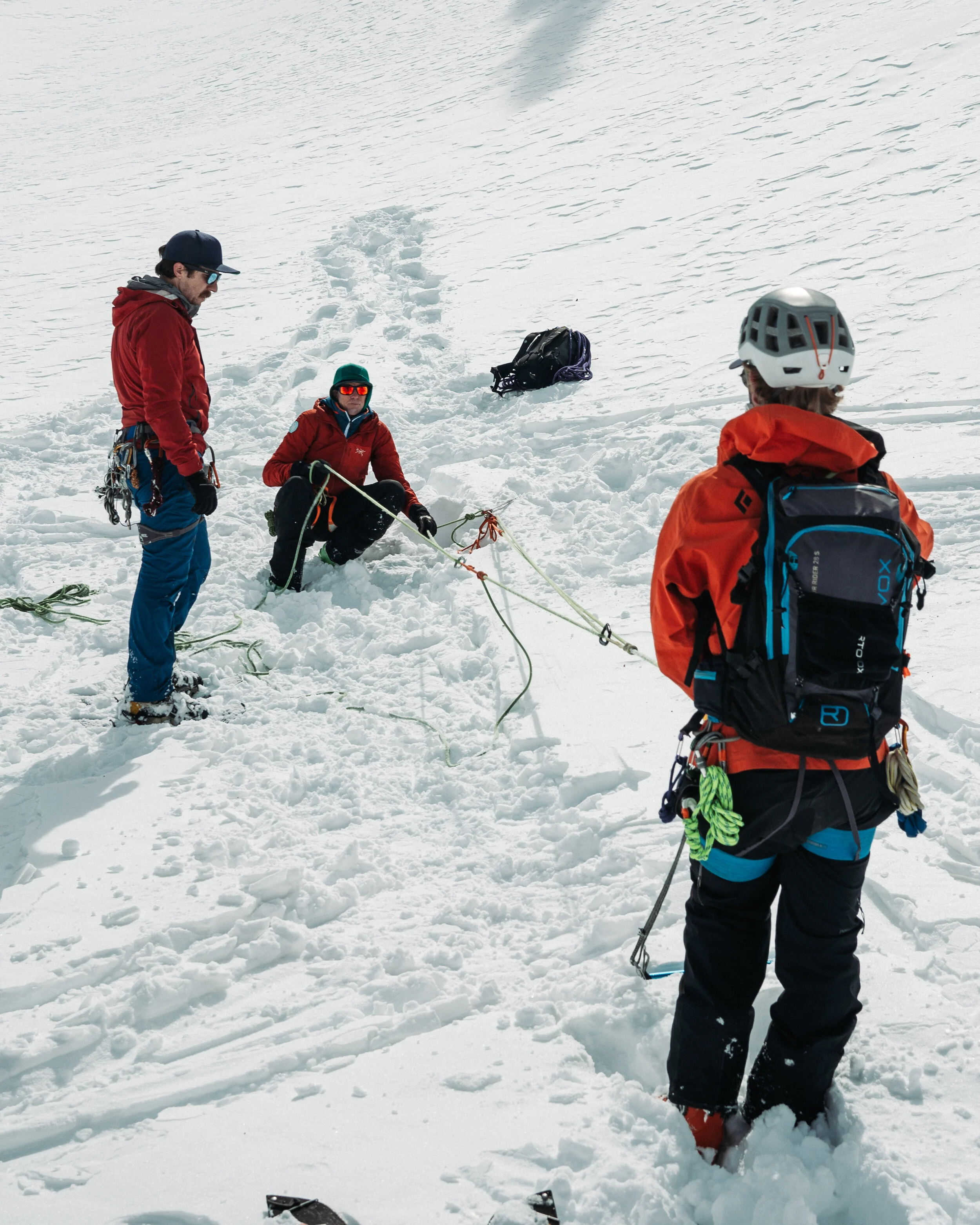 Three mountaineers in snow, one is kneeling and securing a rope, others are standing nearby with gear and backpacks.
