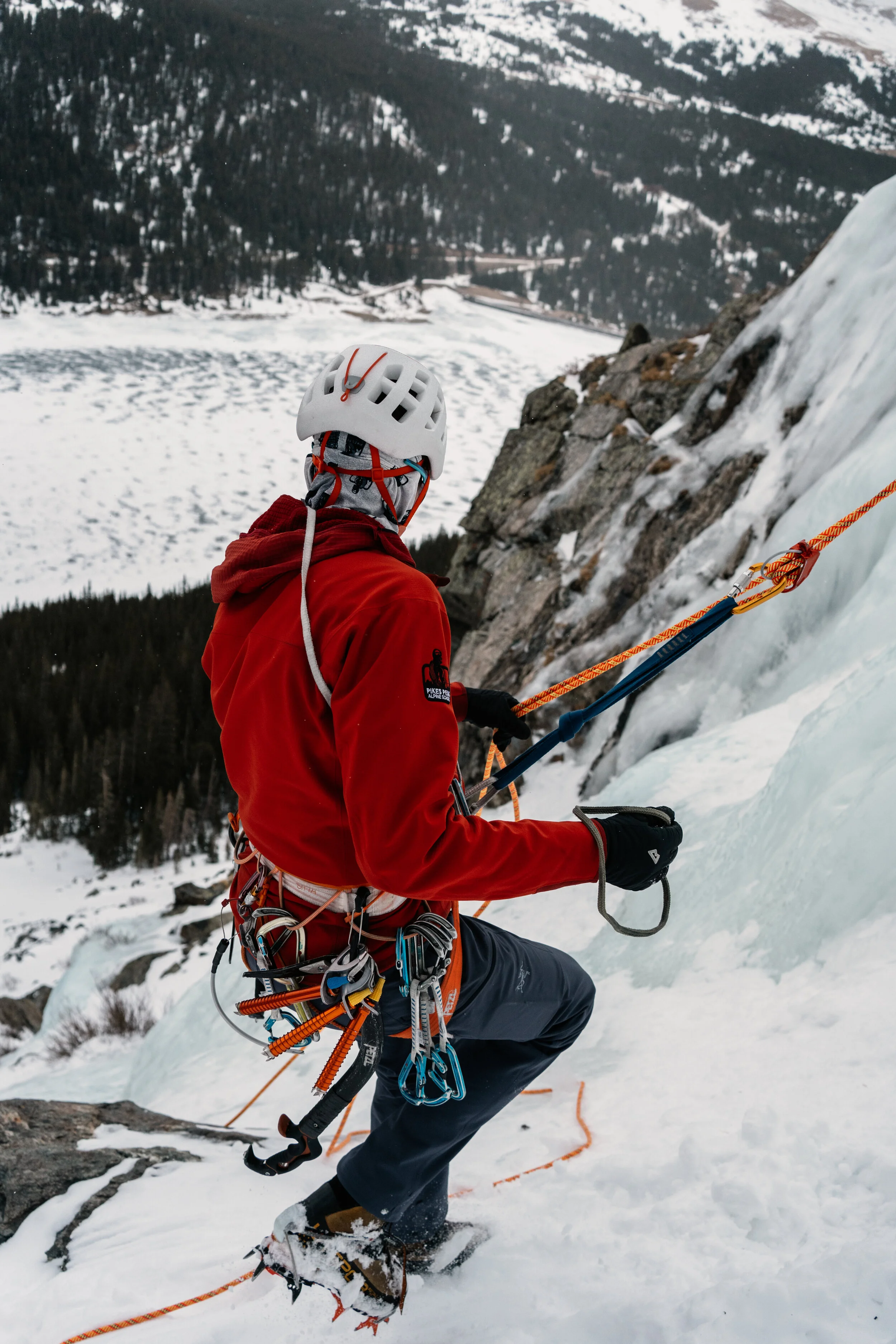 Mountaineer dressed in red jacket and helmet, ascending icy mountain slope using ice axes and ropes.