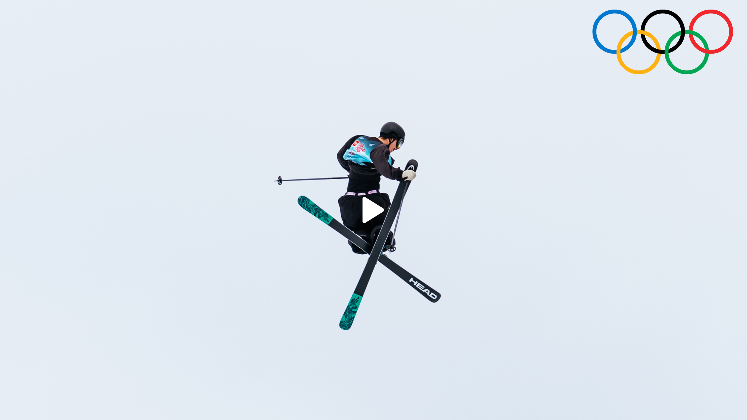A skier performing an aerial trick during the Olympics, with the Olympic rings logo in the top right corner, against a light blue sky.