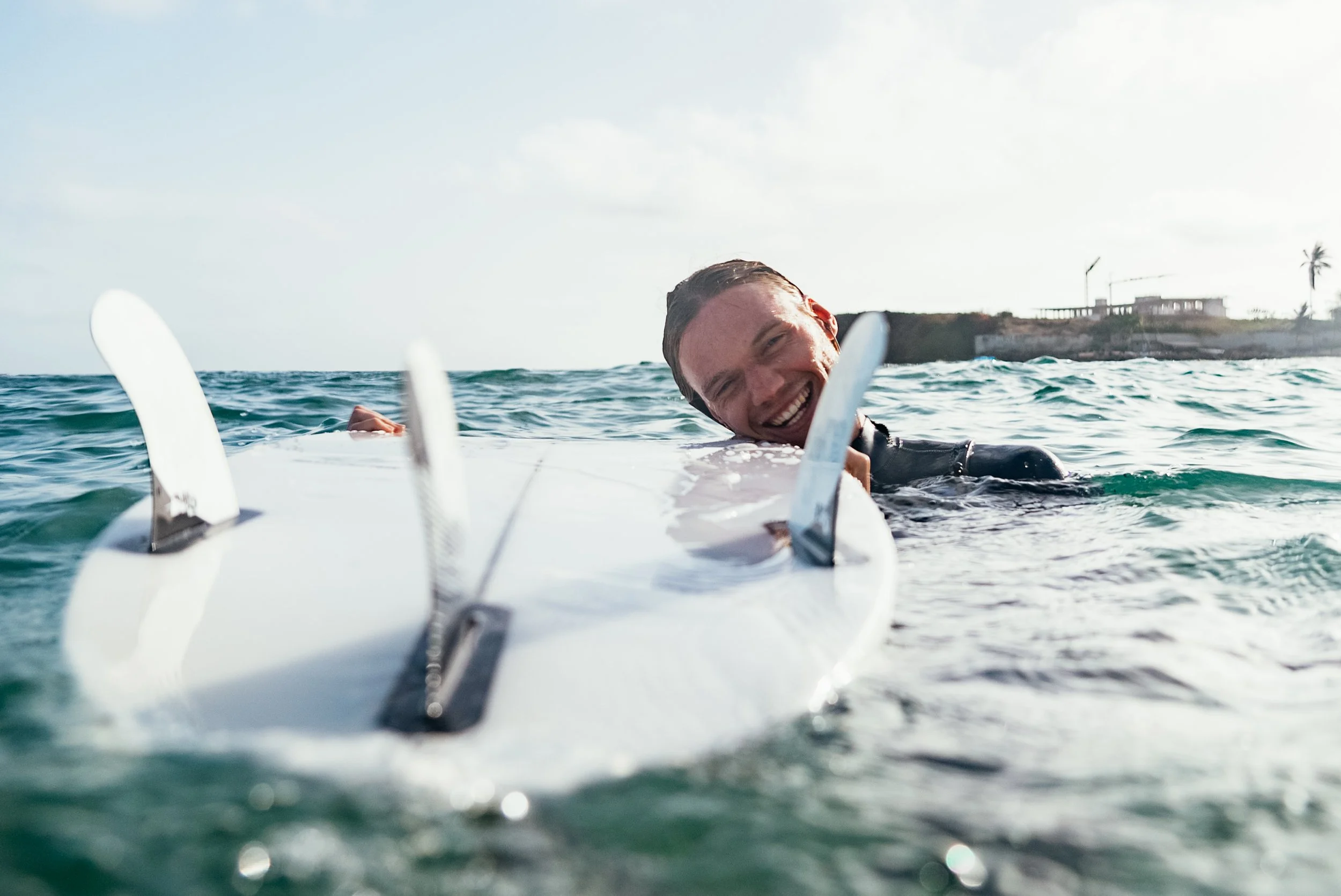 A smiling man in a wetsuit lying on a surfboard in the ocean, with a distant shoreline and some buildings in the background.