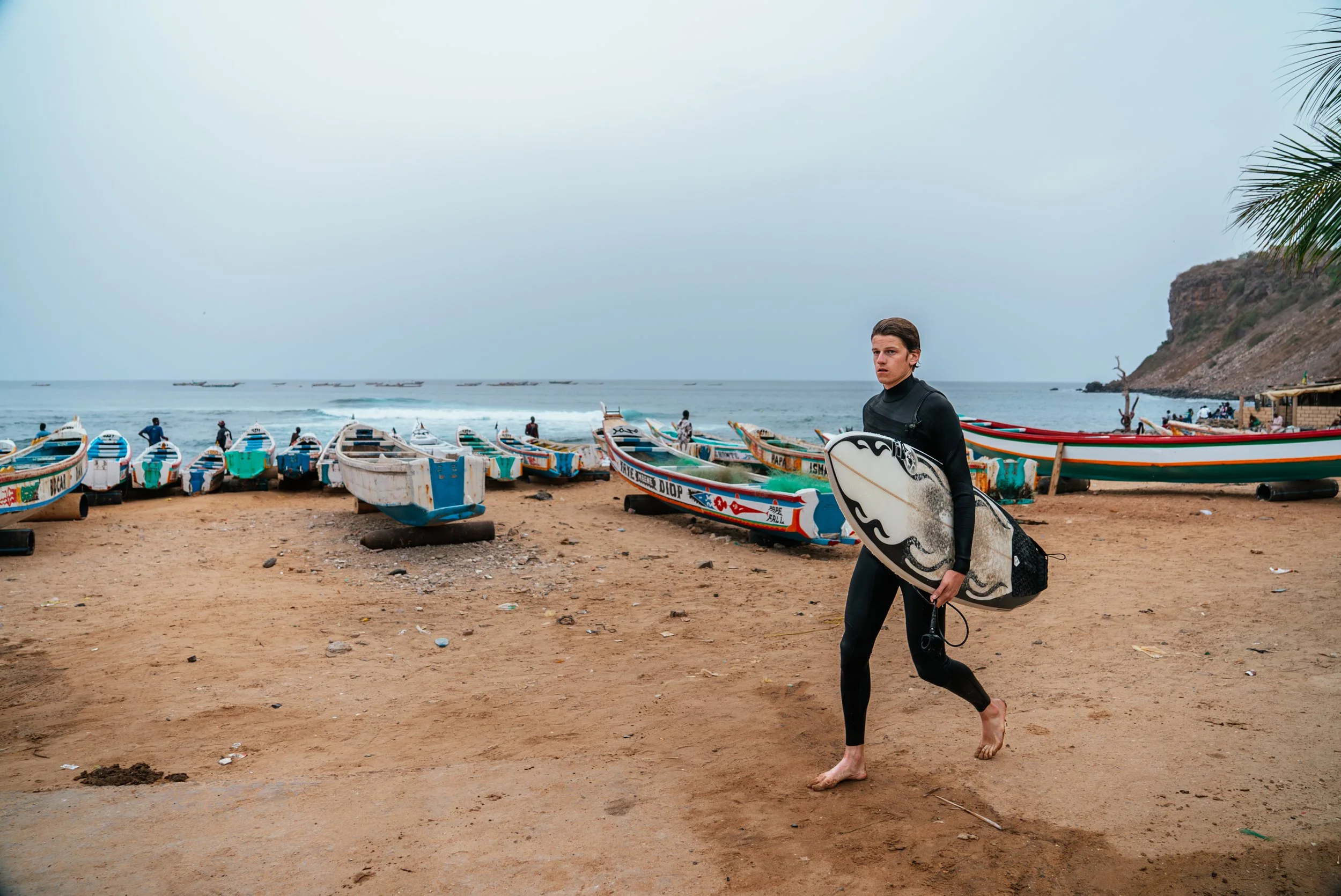 A young man in black wetsuit carrying a surfboard walking on a sandy beach with several colorful boats and a rocky hill in the background.