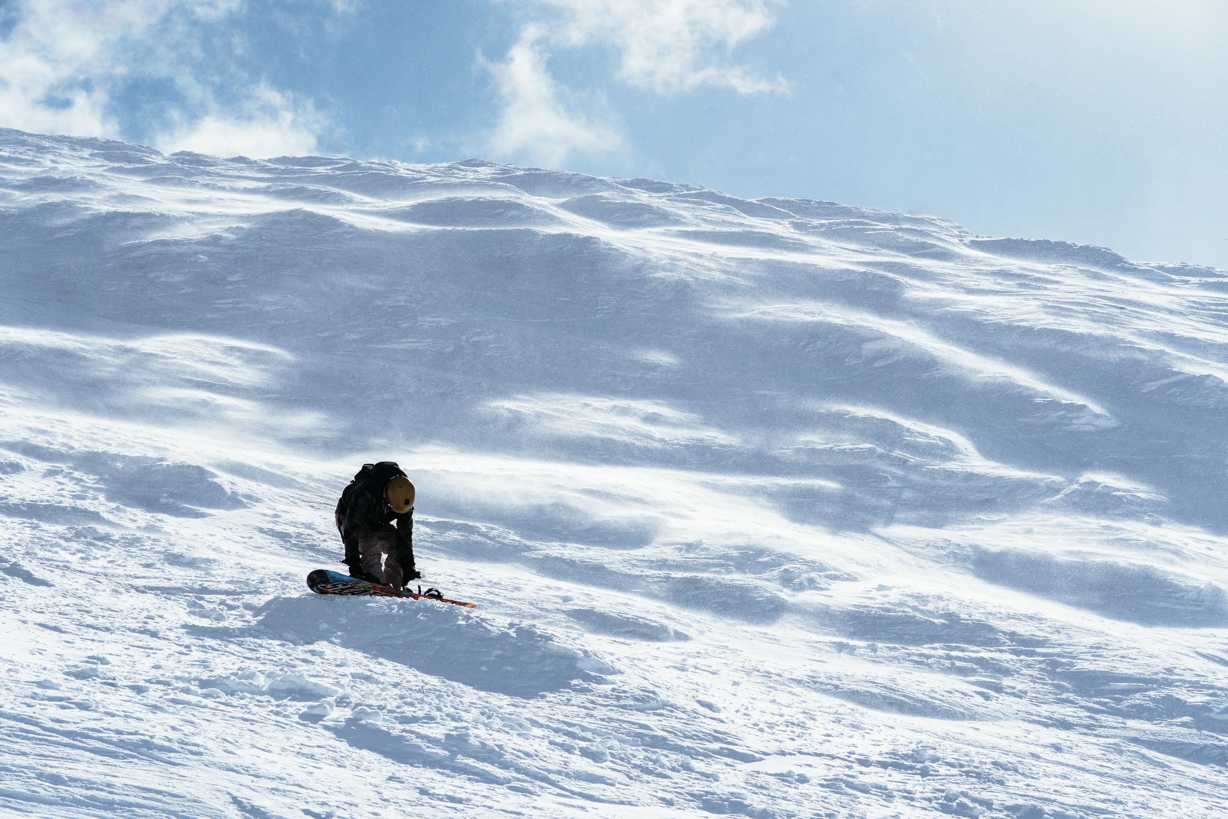 A person wearing gear and a helmet snowboarding down a snowy mountain slope with clear sky and clouds in the background.