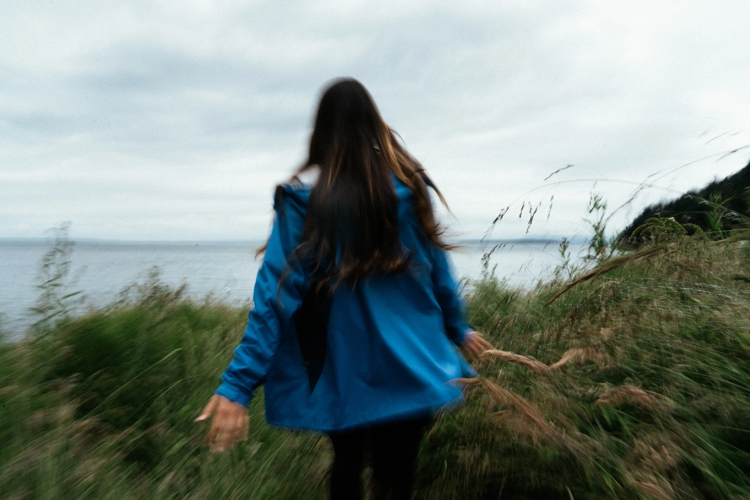 A person with long brown hair wearing a blue jacket walking through tall grass towards a body of water under an overcast sky.