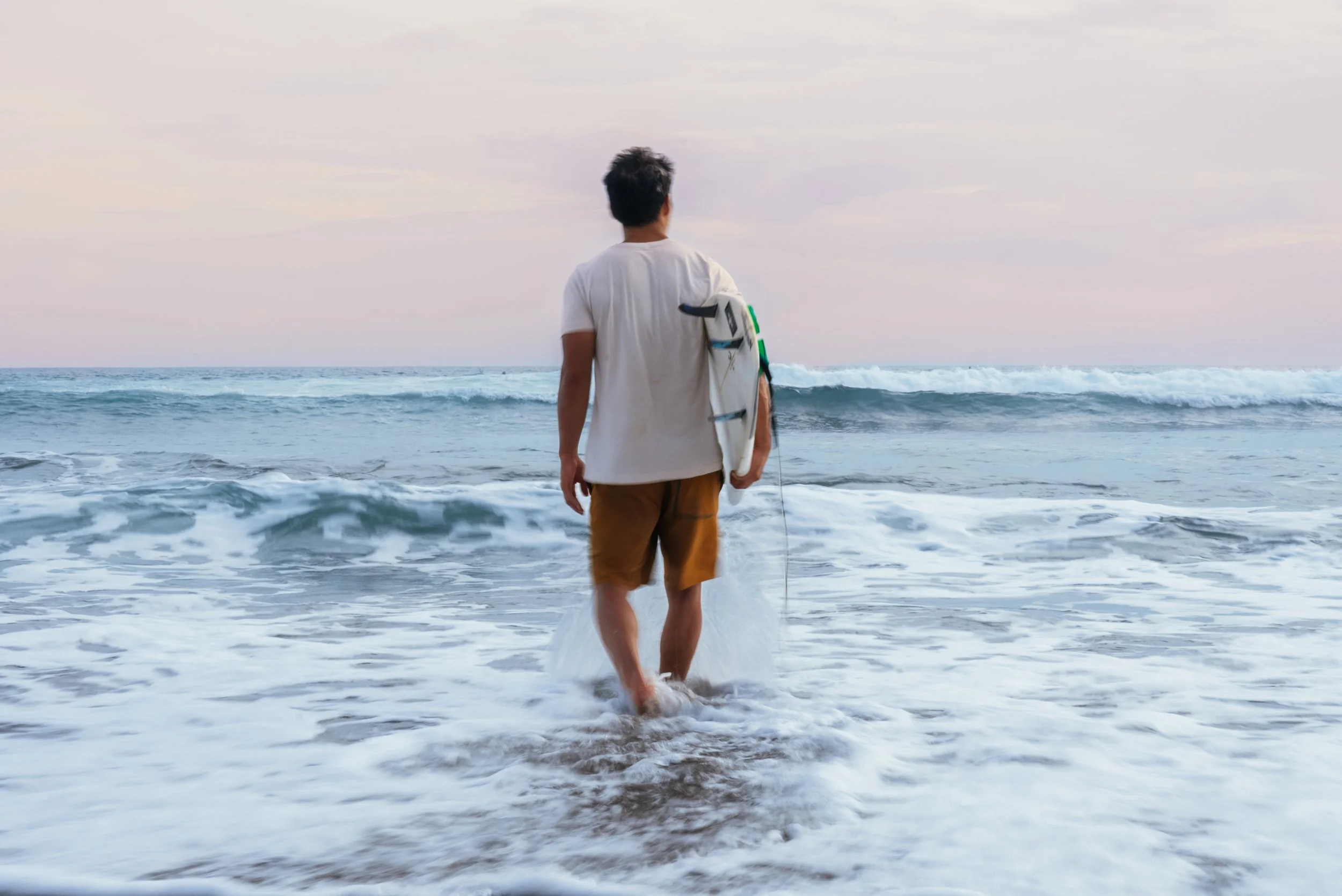 A man in a white t-shirt and brown shorts walking into the ocean while carrying a surfboard under his arm.
