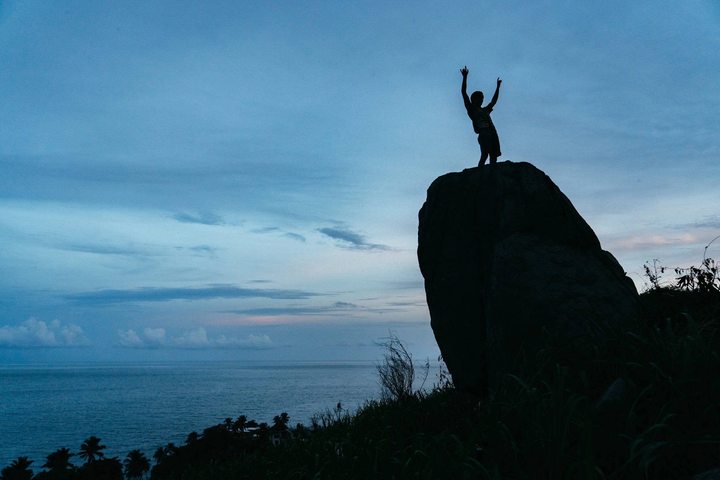 Silhouette of a child standing on a large rock with arms raised, overlooking the ocean during twilight, with some palm trees in the distance.
