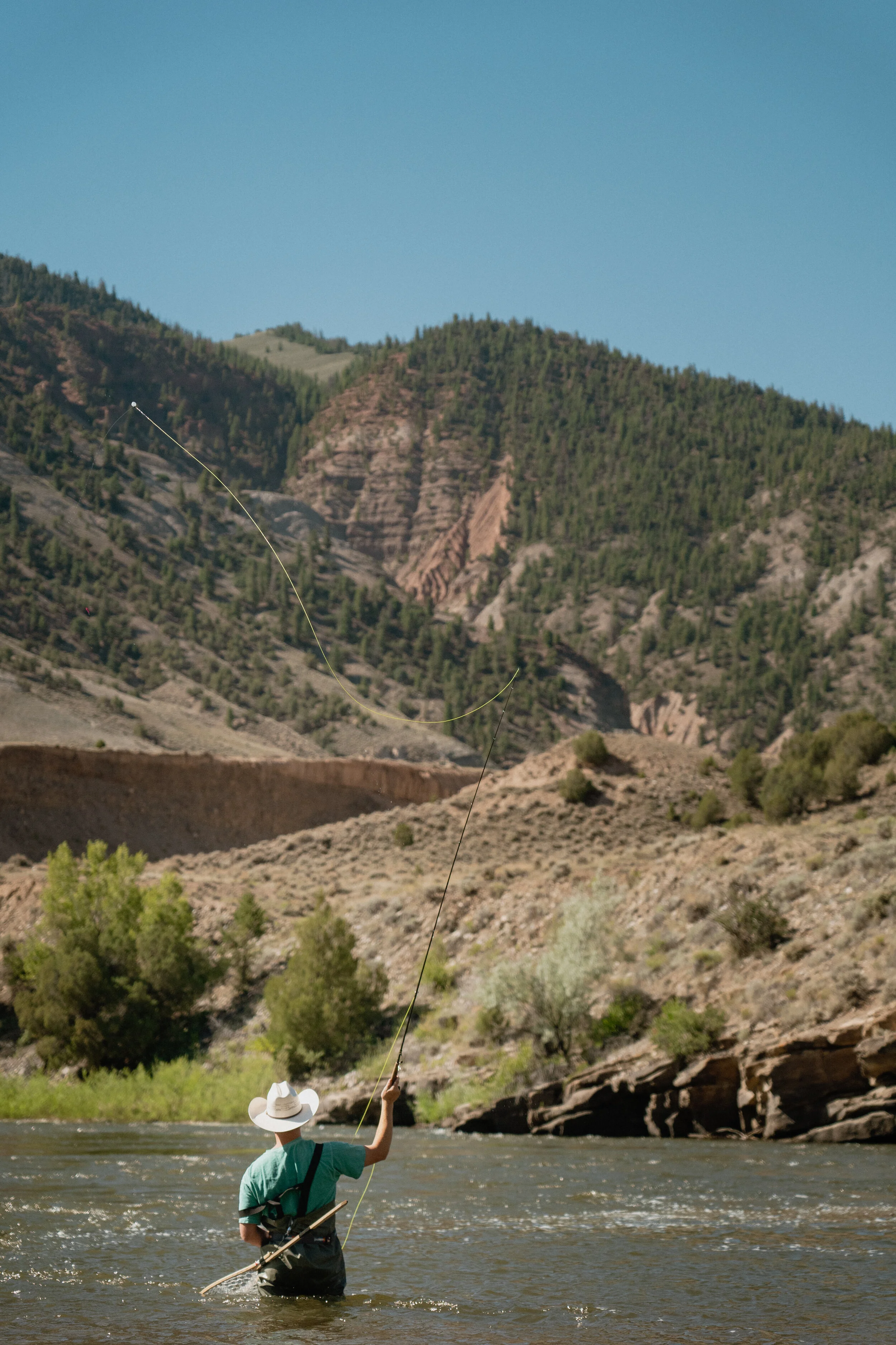 Person fishing in a river with mountains in the background.