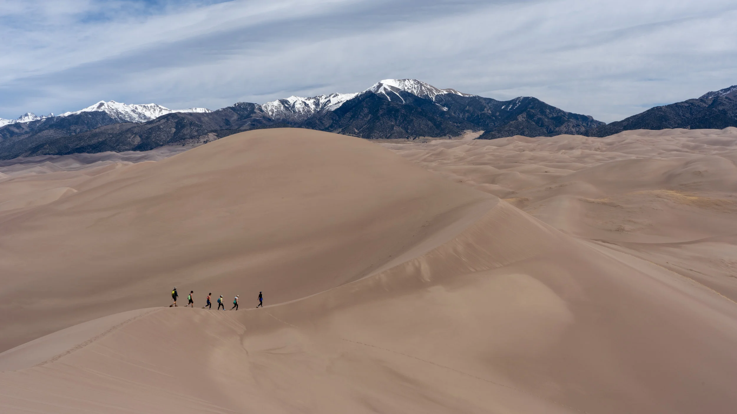 A group of hikers walking across expansive sand dunes with snow-capped mountains in the background under a partly cloudy sky.