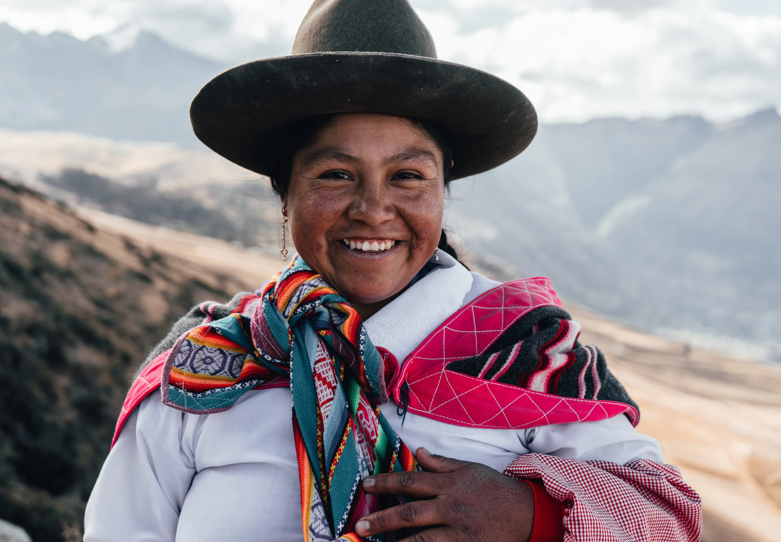 Smiling woman wearing traditional colorful clothing, a black hat, and a patterned shawl outdoors with a mountainous landscape in the background.