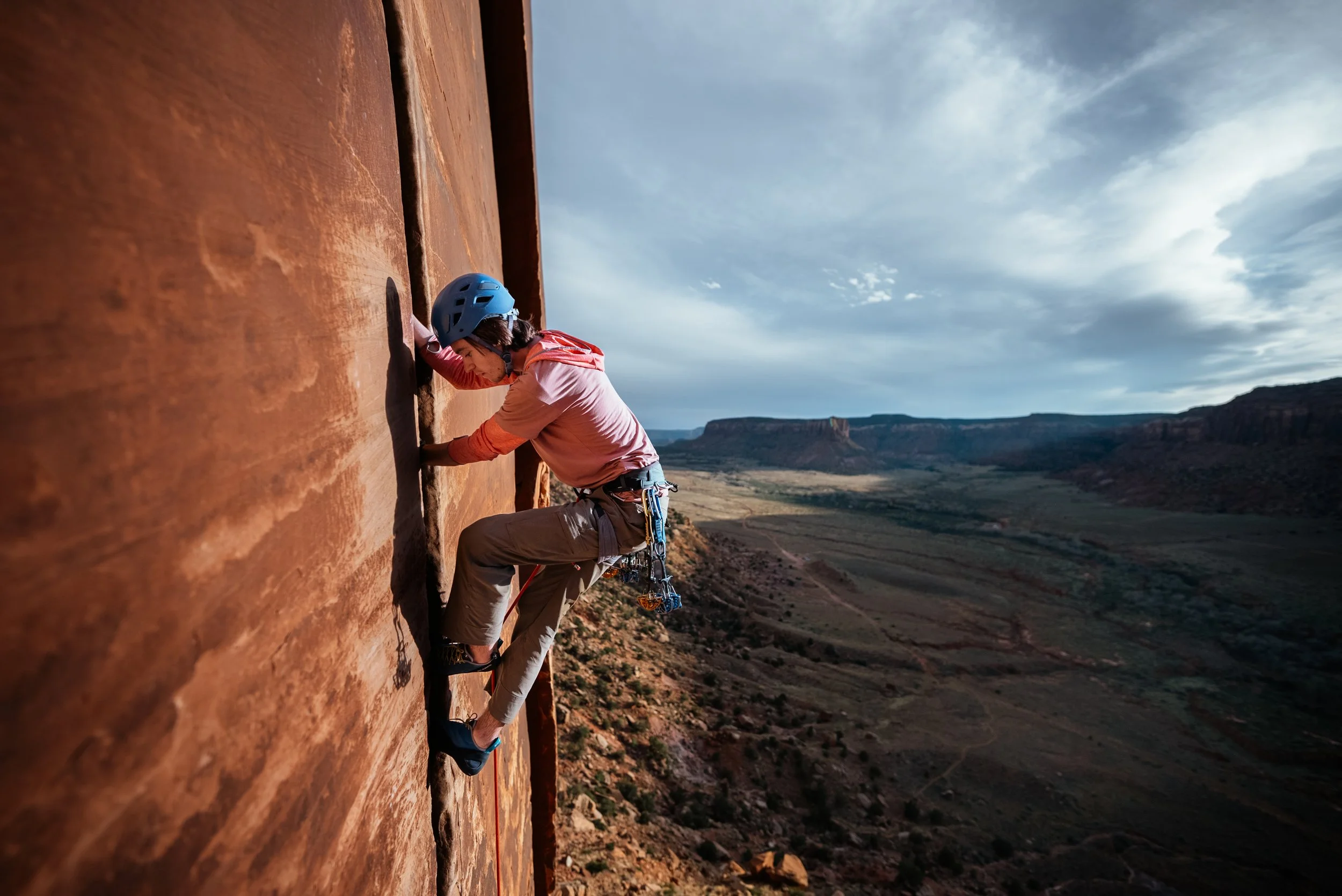A person in a pink shirt and tan pants rock climbing a steep red sandstone cliff with a helmet and climbing gear, overlooking a desert canyon landscape under a cloudy sky.