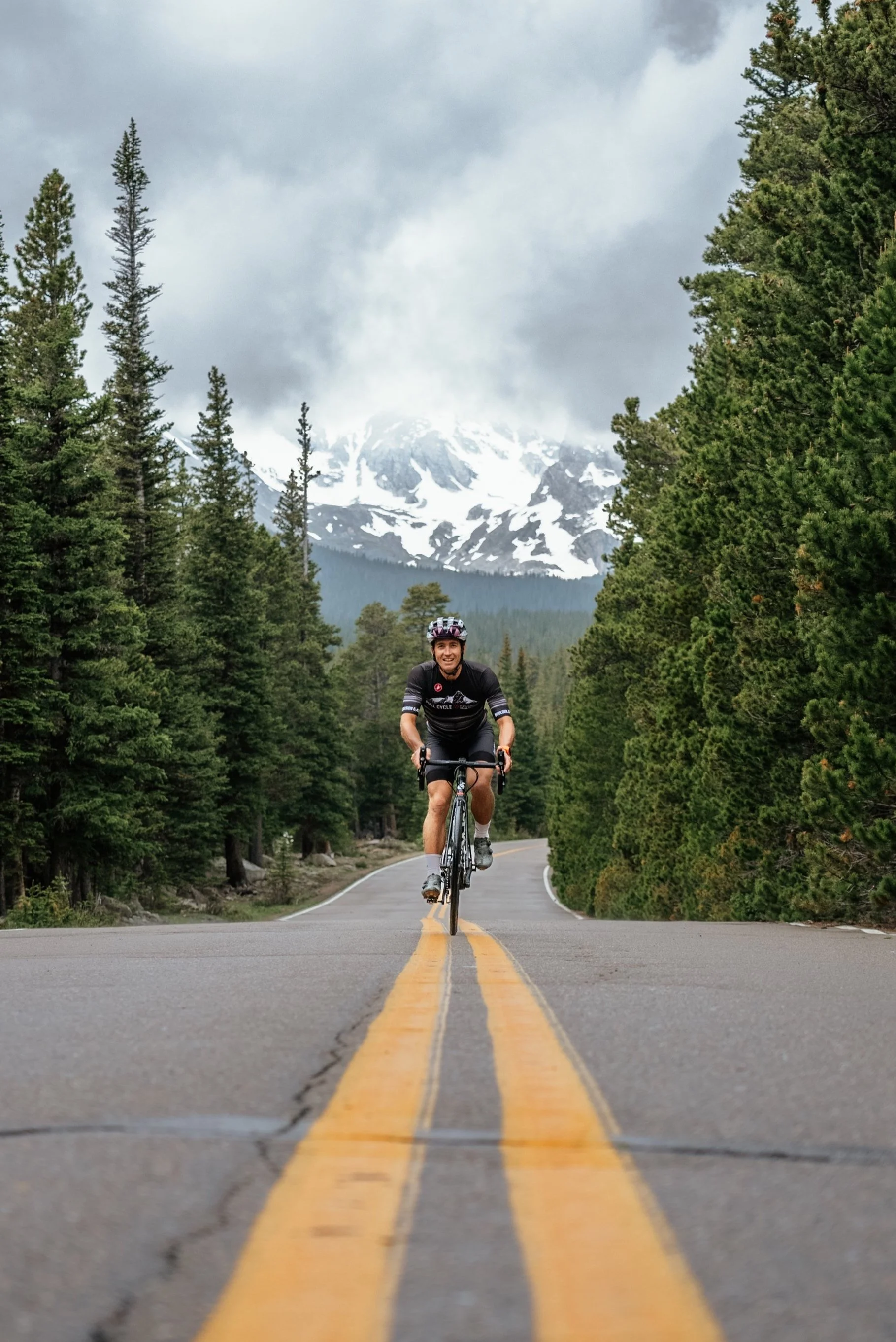 A man riding a bicycle on a mountain road surrounded by tall pine trees, with snow-capped mountains and cloudy sky in the background.