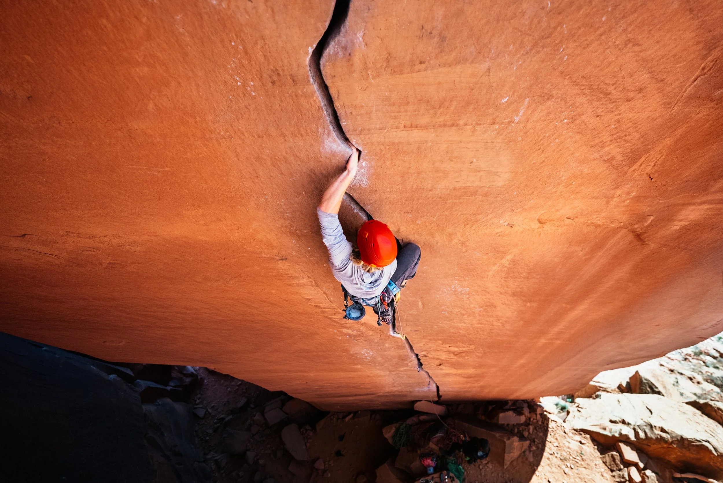 A rock climber in an orange helmet climbing a tall, reddish, vertical rock face in a canyon.