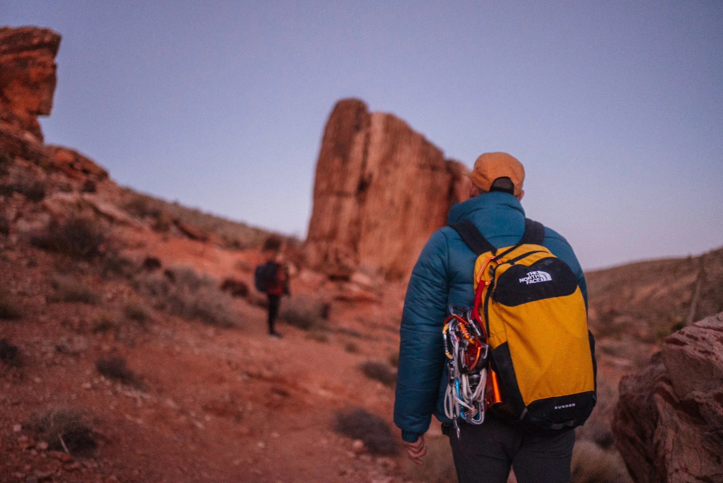 Two hikers with backpacks walking on a rocky trail through a desert landscape at dawn or dusk, with large rock formations and a clear sky in the background.