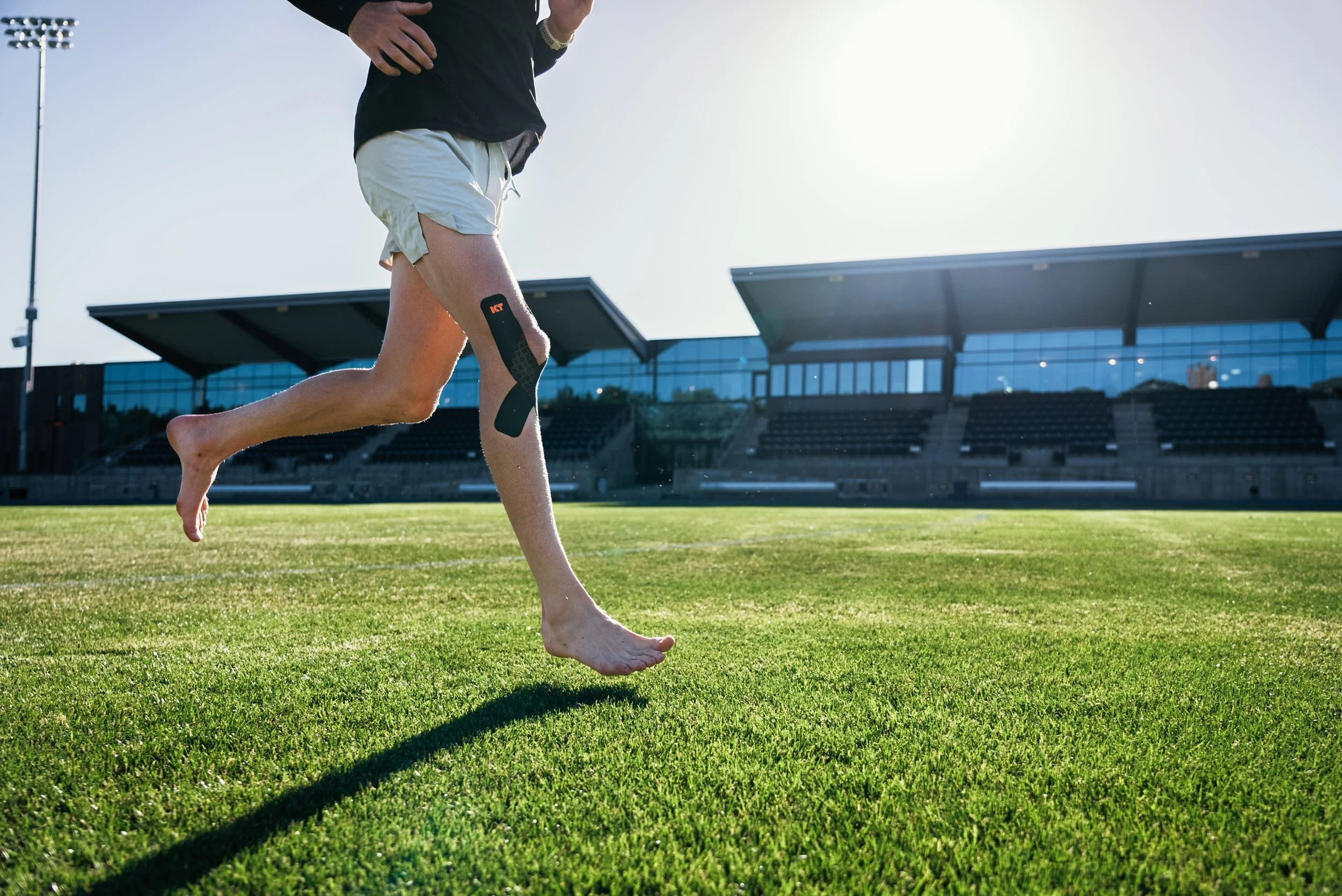 A person running barefoot on a grassy field, wearing a prosthetic leg with a black and orange attachment, in an outdoor stadium with modern glass buildings in the background.